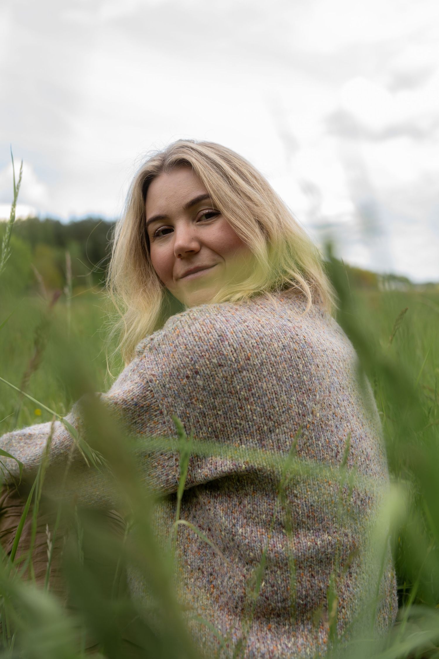 Smiling woman with long blonde hair in a knit sweater sits in a green grassy field under a cloudy sky.