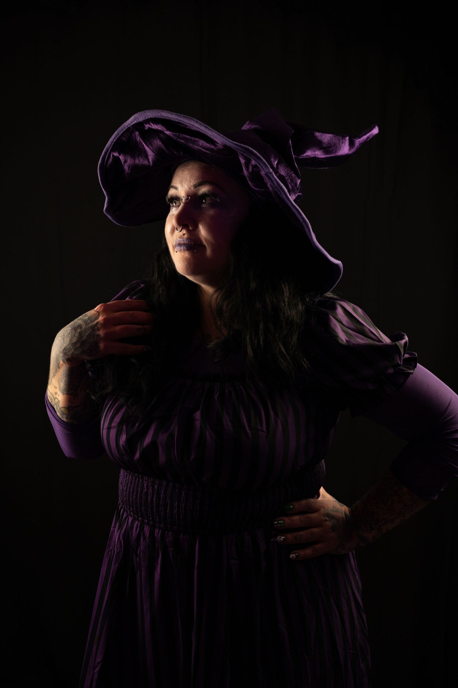 A woman in a purple witch hat and striped dress poses dramatically against a dark background in a moody Halloween-style portrait.