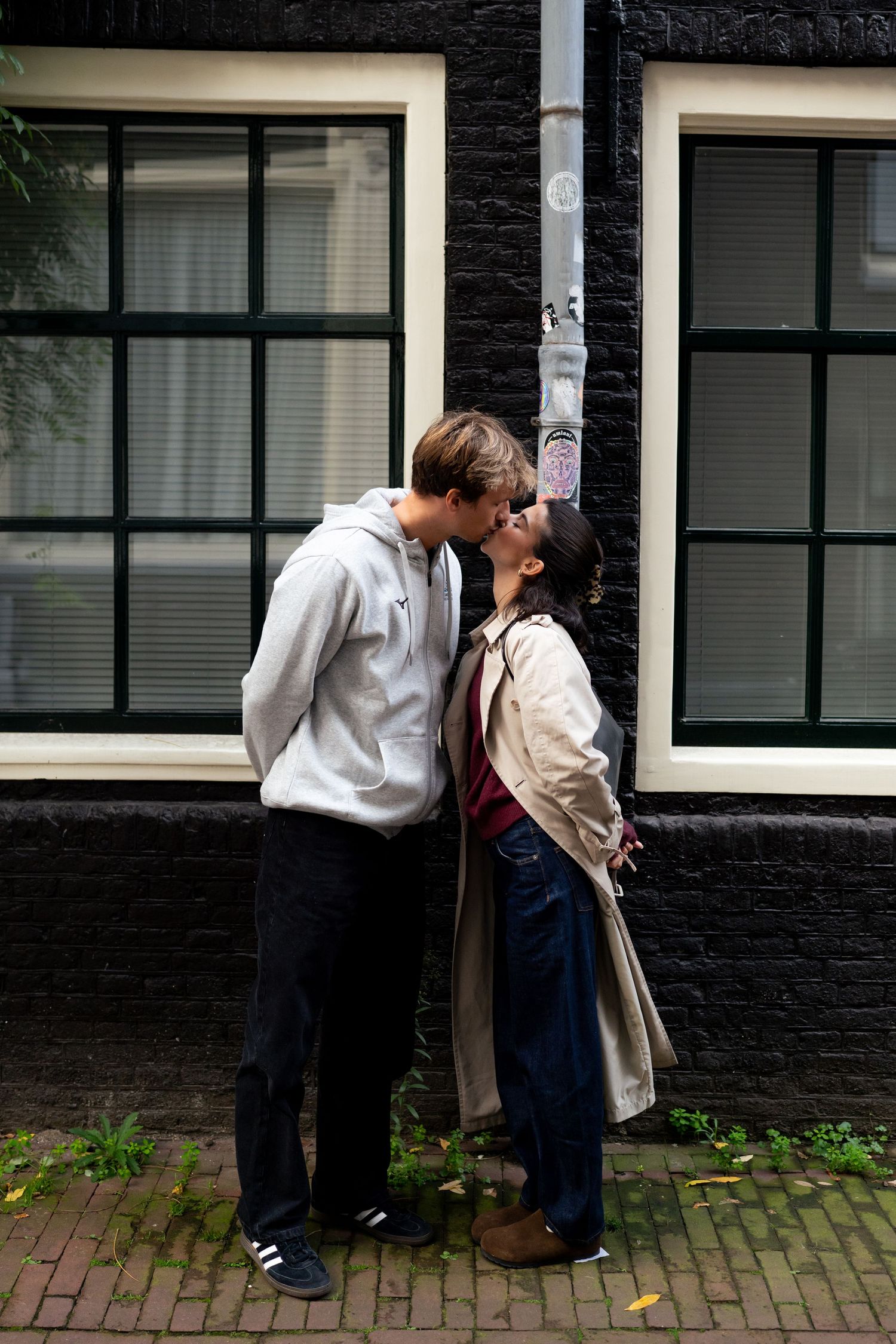 Couple kissing on a brick sidewalk in front of a dark brick building with large white-framed windows.