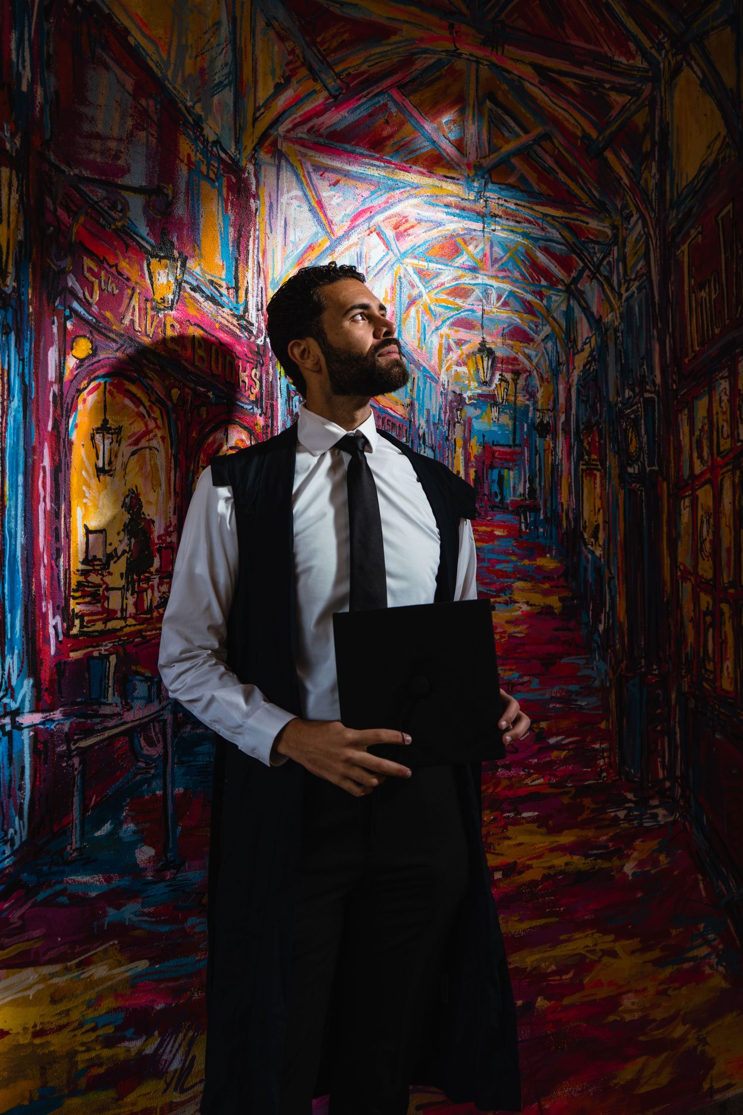 Bearded man in white shirt, black tie and black academic gown stands in a vividly painted, colorful corridor with an arched vaulted ceiling.