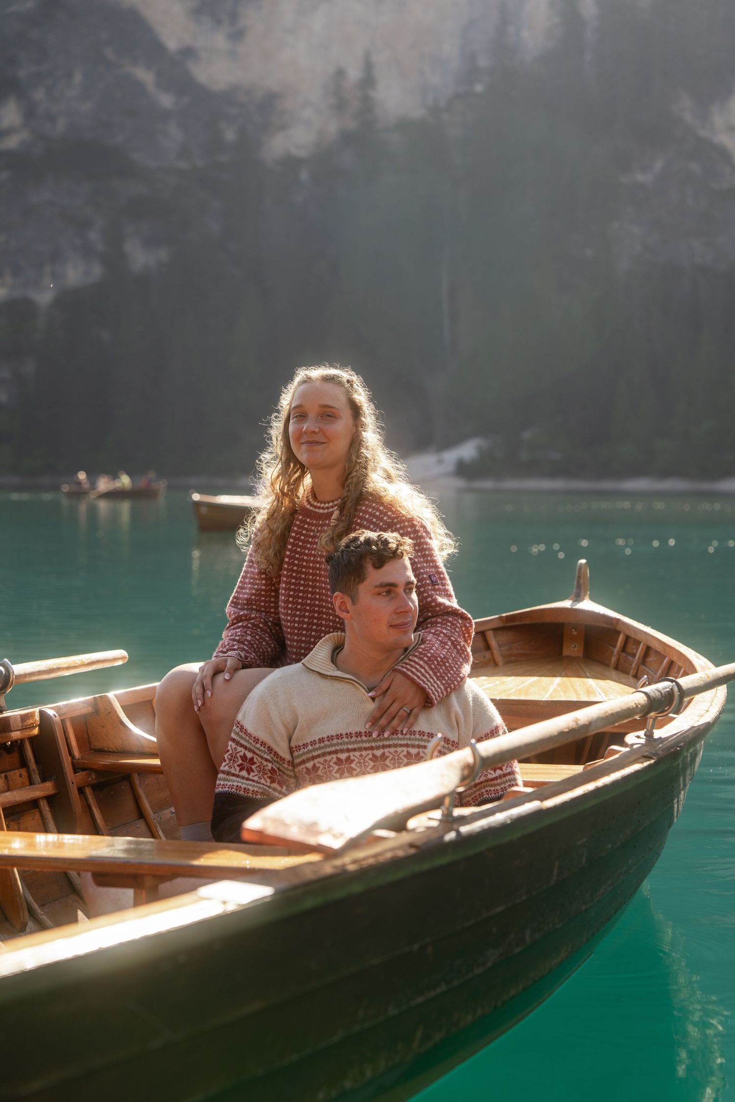 A couple in matching patterned sweaters sits together in a wooden rowboat on a turquoise mountain lake surrounded by evergreen forest.