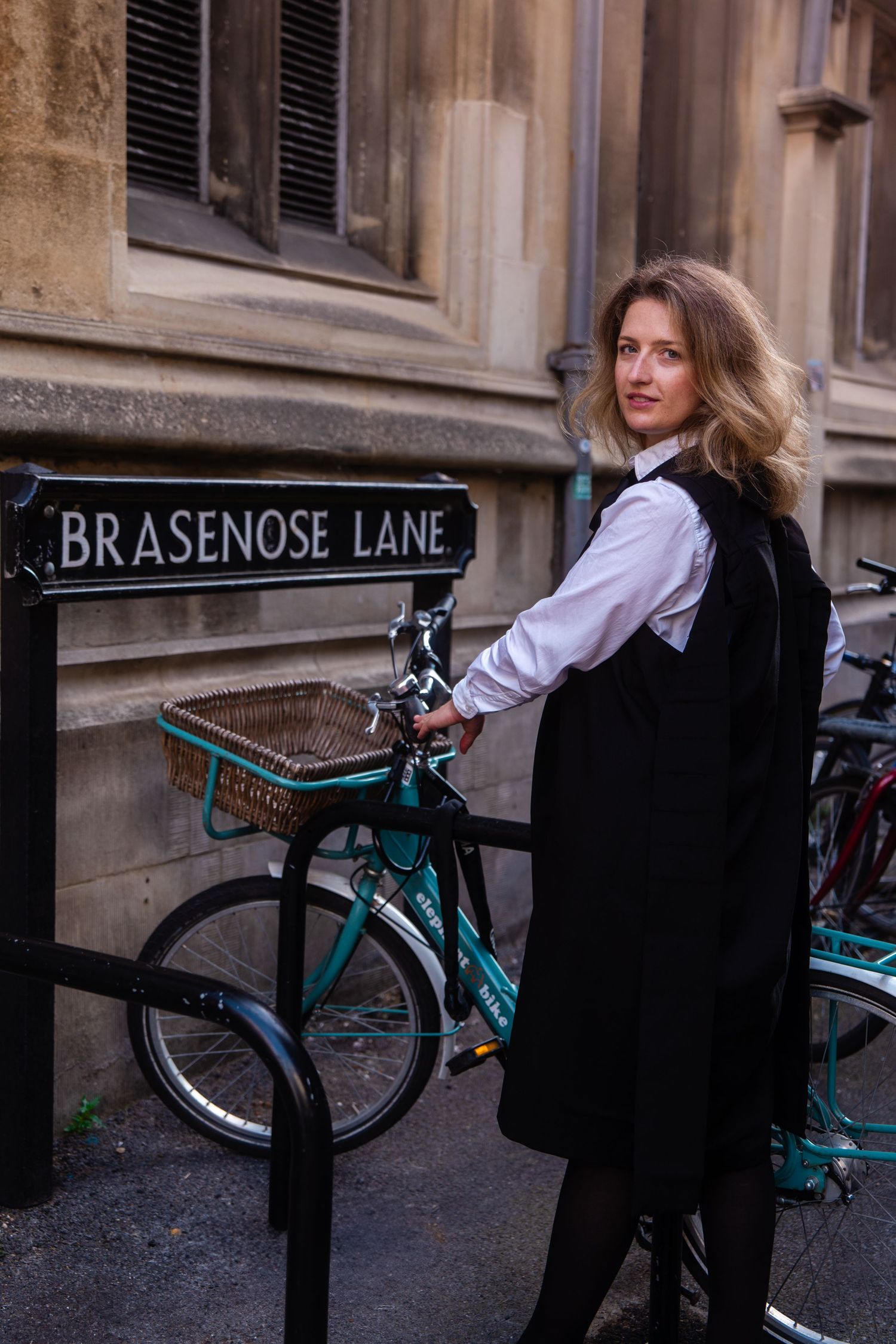 Woman in a white shirt and black pinafore dress stands beside a teal bicycle with a wicker basket next to a Brasenose Lane street sign.