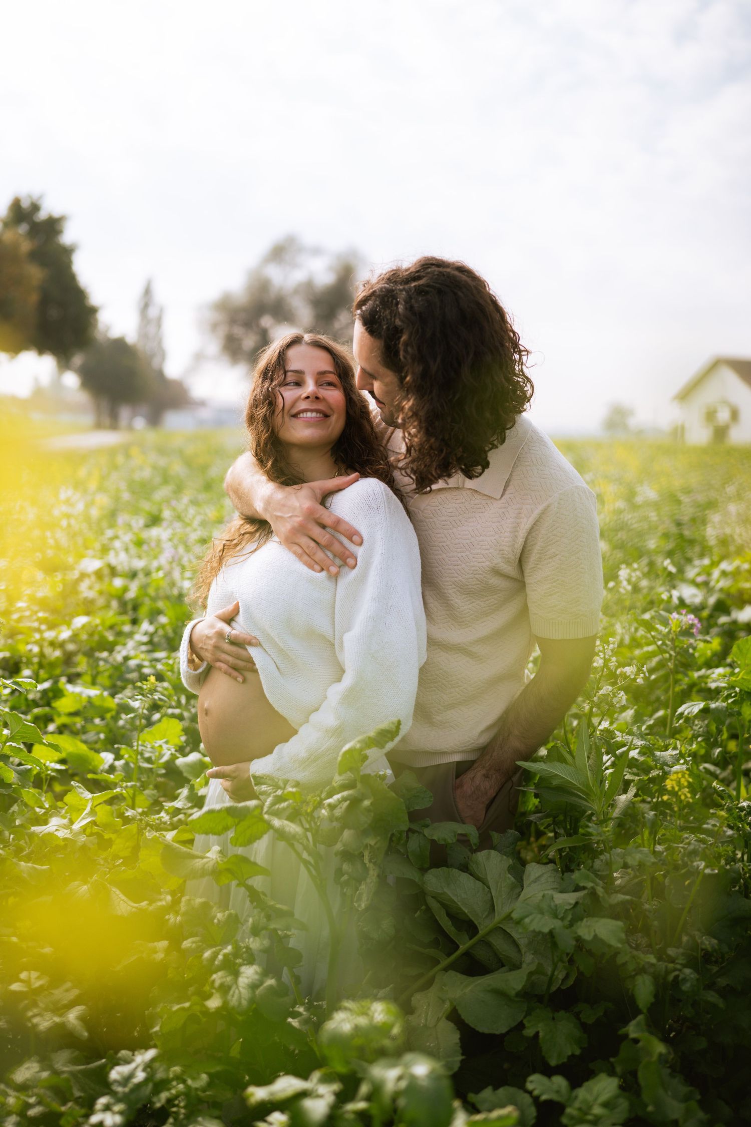 Smiling pregnant woman in a white sweater is embraced from behind by her curly-haired partner in a sunlit green field.