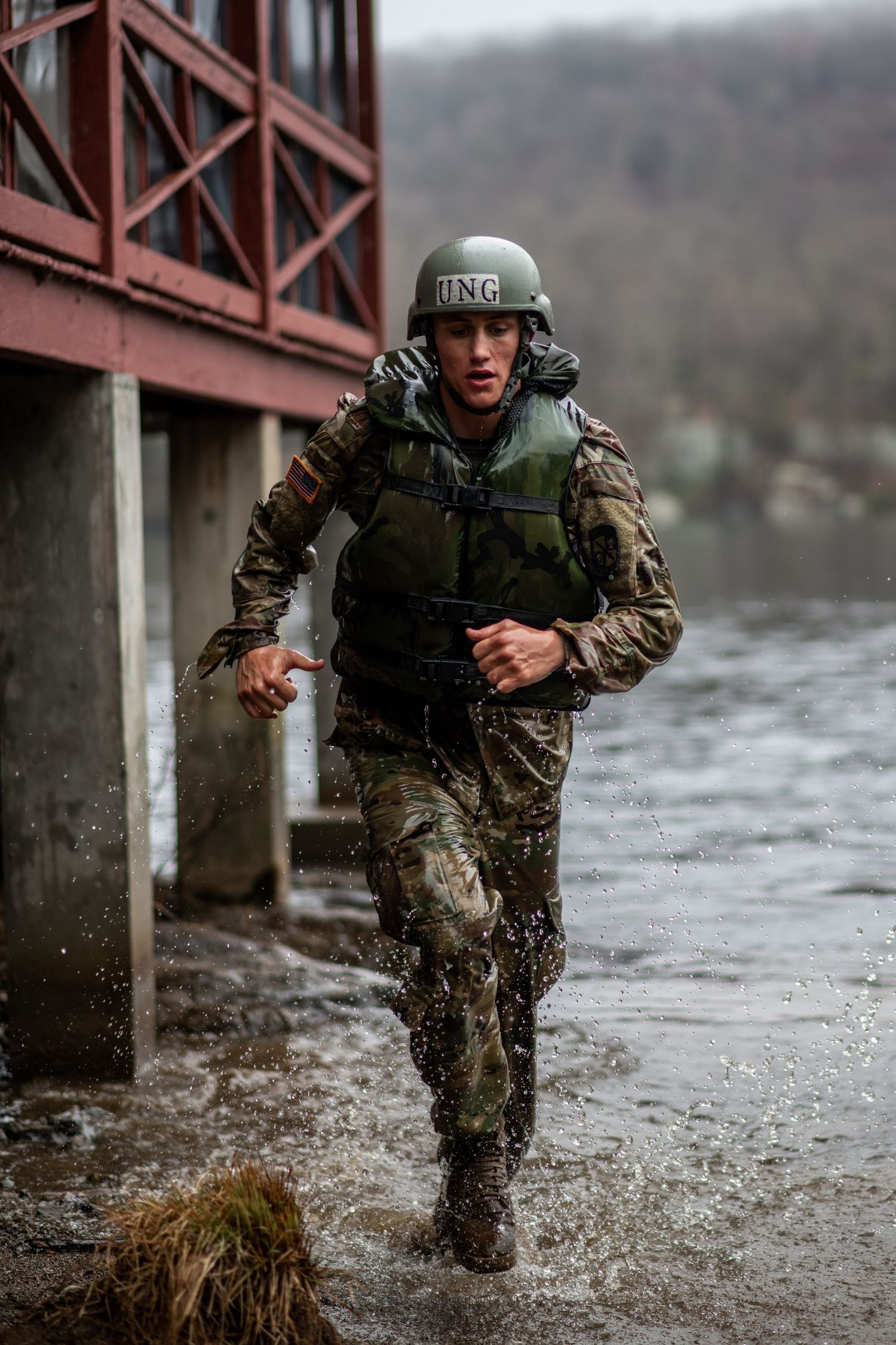 A soldier in camouflage, helmet and tactical vest runs through splashing water beside a red bridge structure.