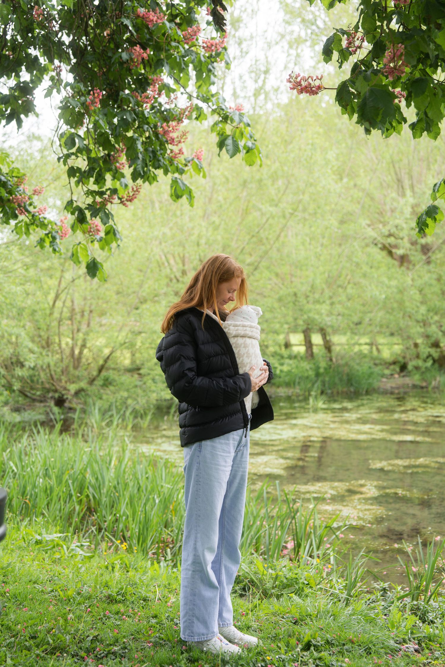 Woman in a black puffer jacket and light jeans cradles a swaddled baby beside a green pond under blooming chestnut trees.