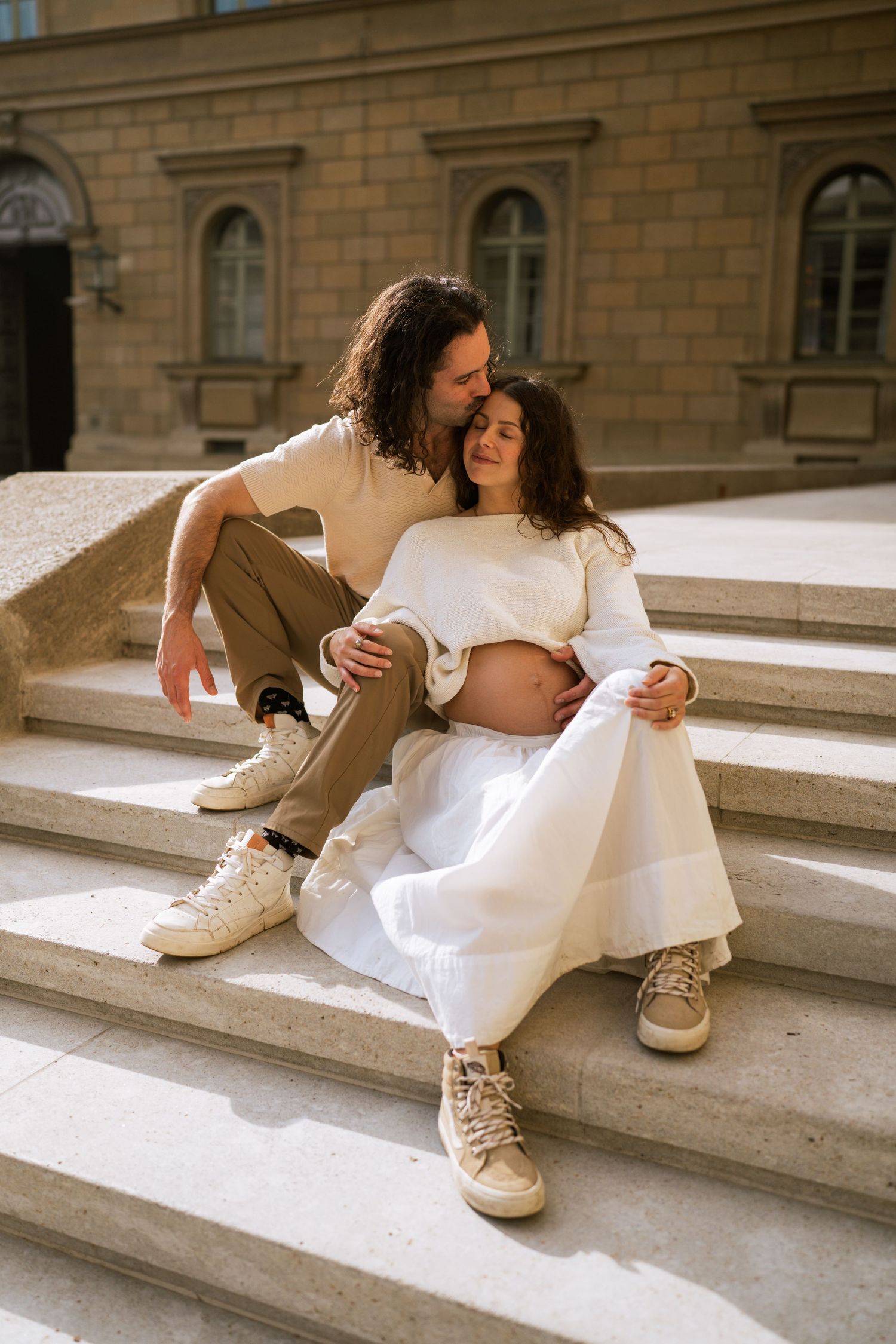 Expecting couple sits close together on stone steps as the man kisses the woman's forehead during an outdoor maternity photoshoot.