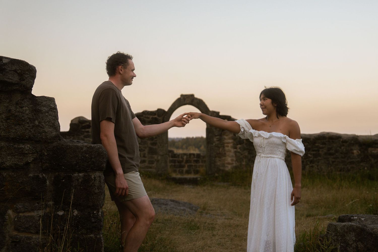 A man and a woman in a white off-the-shoulder dress hold hands among ancient stone ruins at sunset.