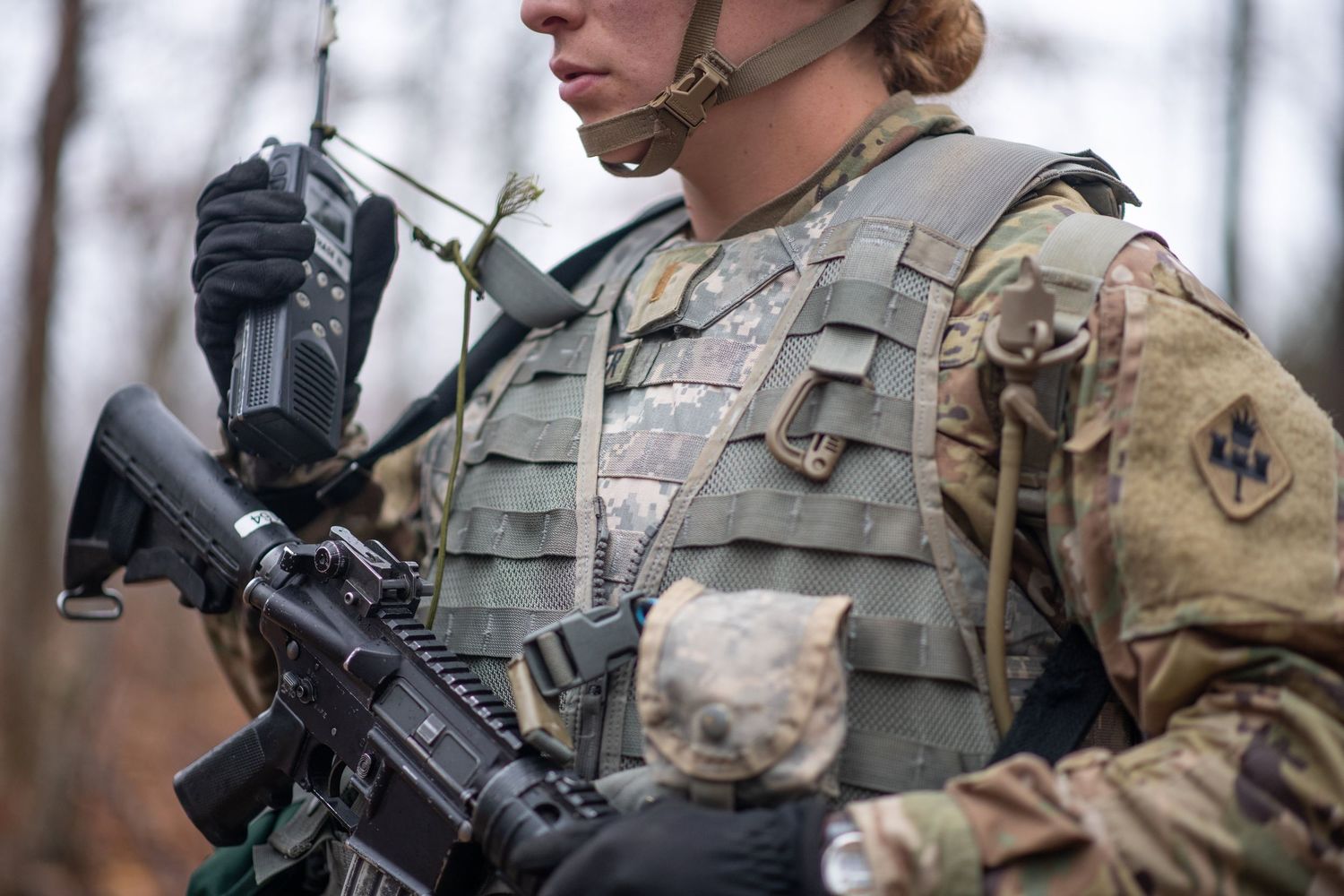 Close-up of a soldier in camouflage and a tactical vest holding a rifle and a radio in a wooded area.
