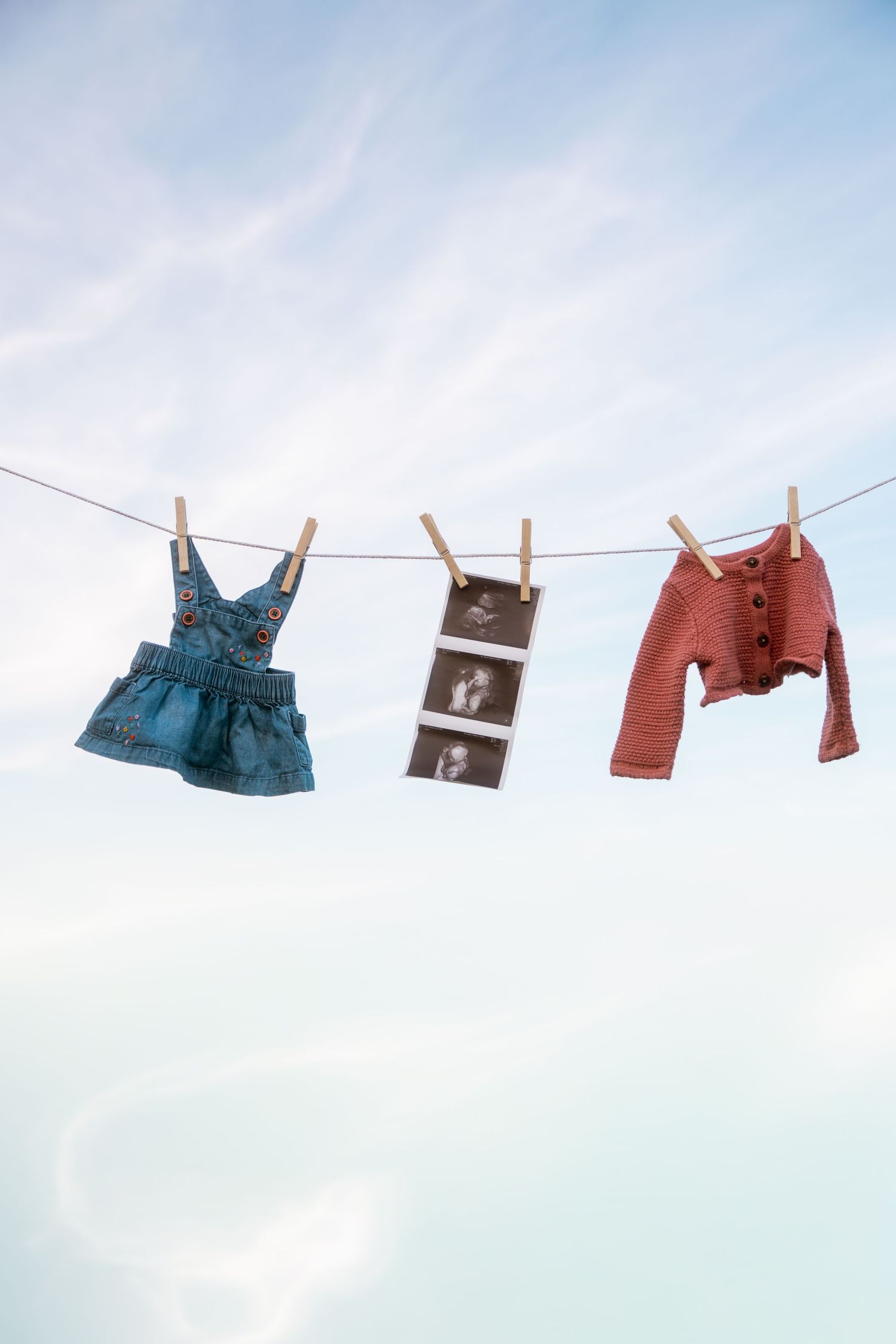 Baby denim dress, ultrasound photos and a small coral cardigan hang from wooden clothespins on a clothesline against a soft blue sky.