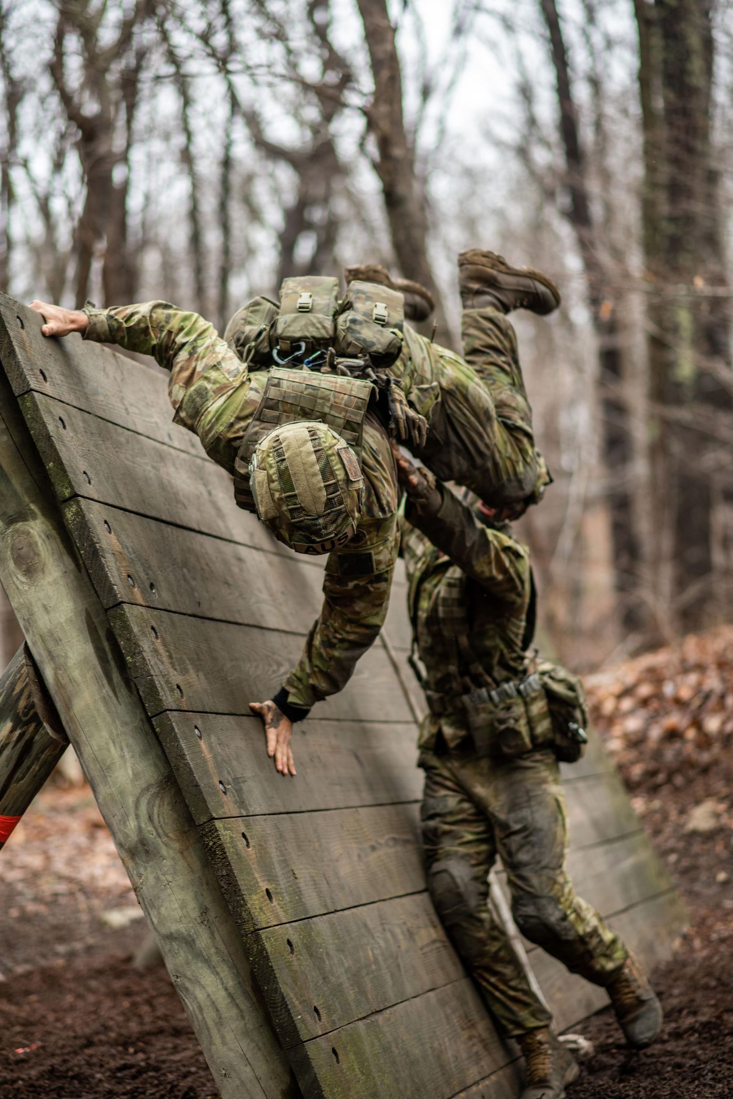 Soldiers in camouflage and helmets climb over a tall wooden wall on a forest obstacle course.