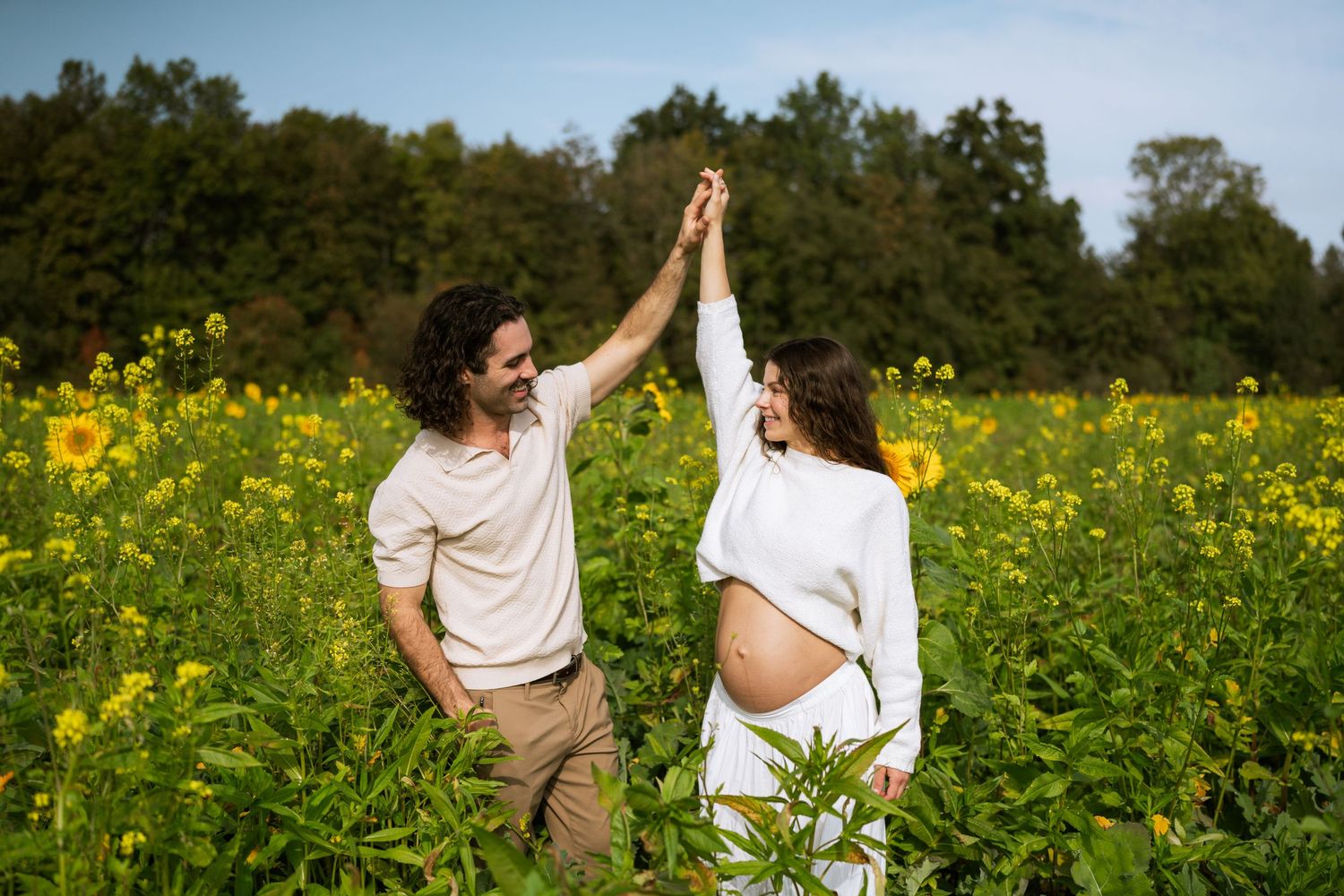 A smiling couple holds hands and dances in a sunflower field during a golden-hour maternity photoshoot with the woman's baby bump visible.