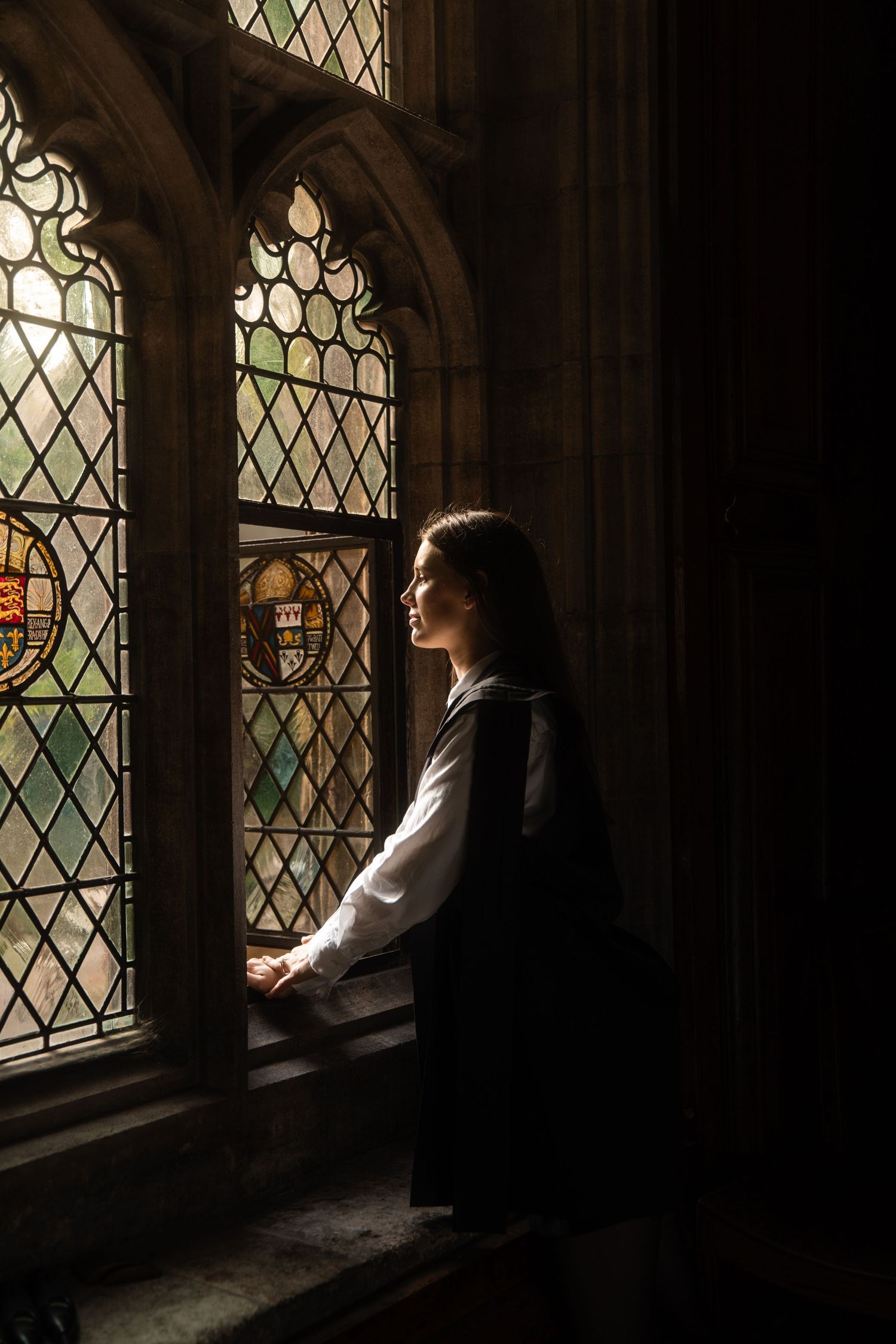 A young woman in a dark academic gown leans on a stone windowsill, gazing through leaded stained glass windows.