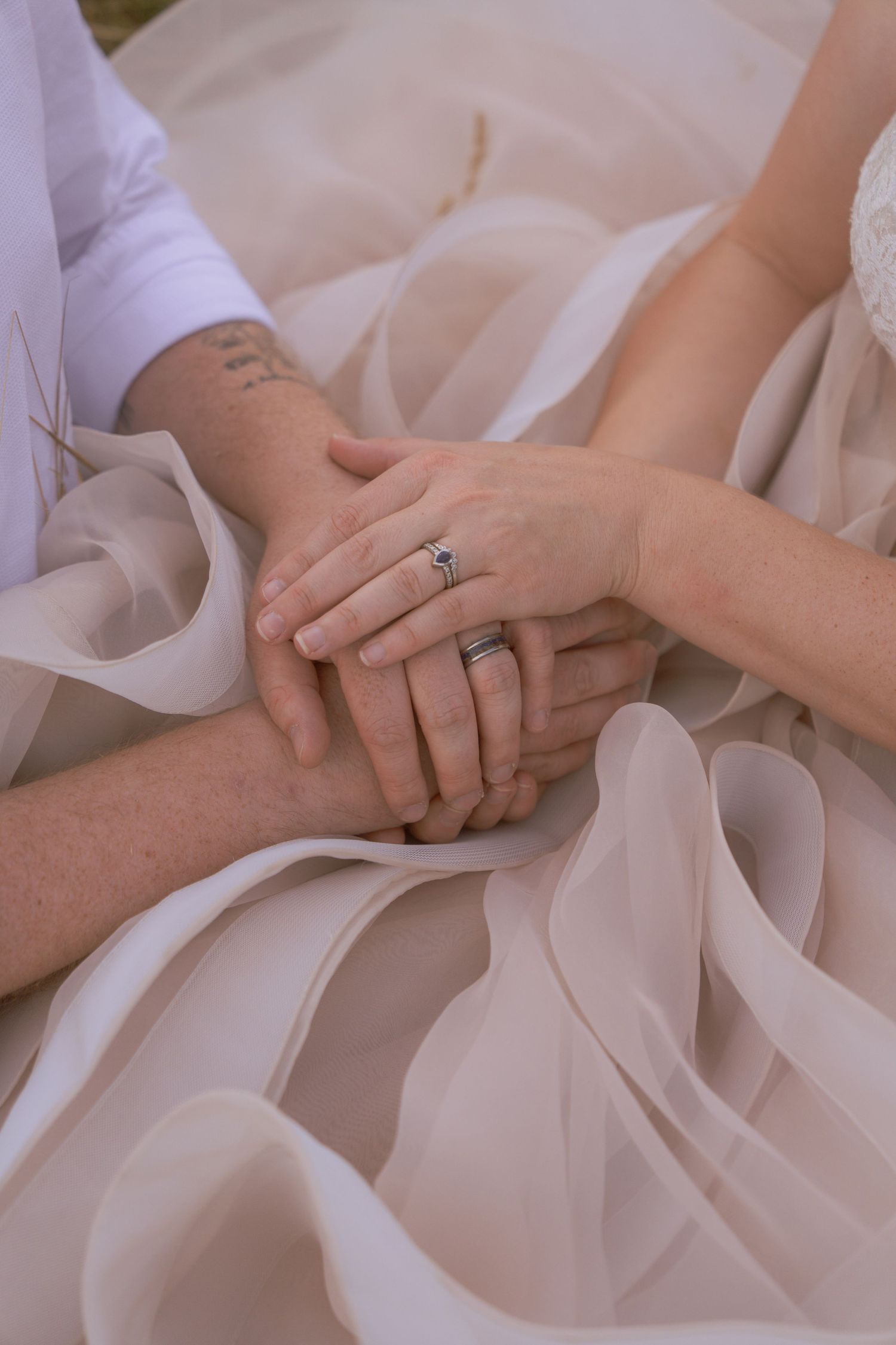 Close-up of a couple's clasped hands wearing wedding rings, resting on a flowing blush tulle wedding dress.