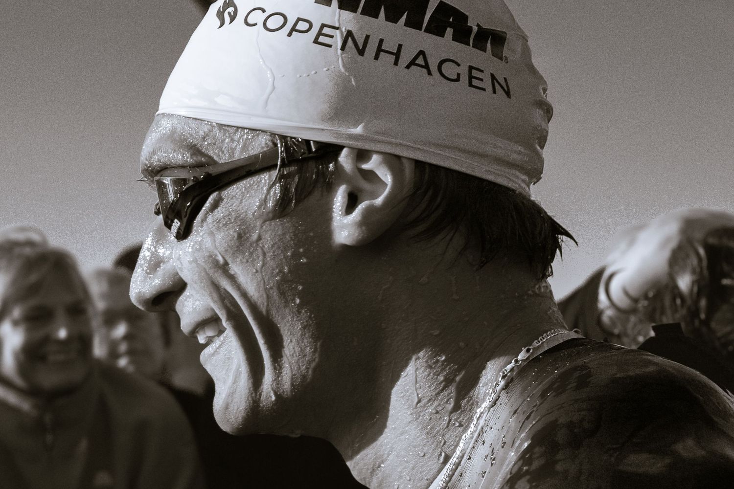 Black and white profile of a smiling athlete in a Copenhagen swim cap and goggles, wet skin glistening, with spectators in the background.