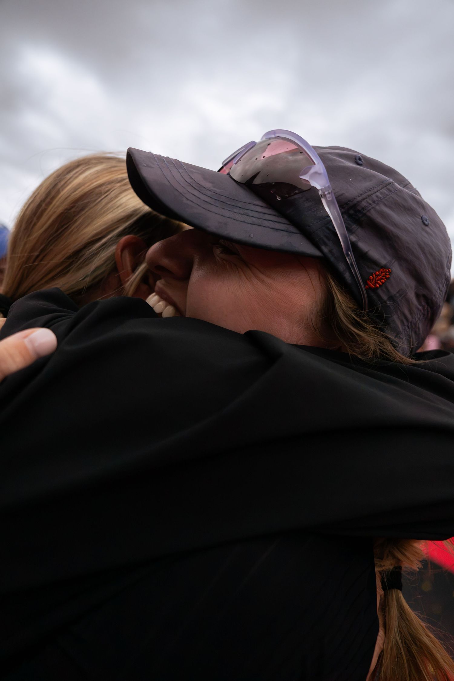 Two people share an emotional hug under a cloudy sky, one wearing a gray baseball cap with sunglasses resting on top and a black jacket.