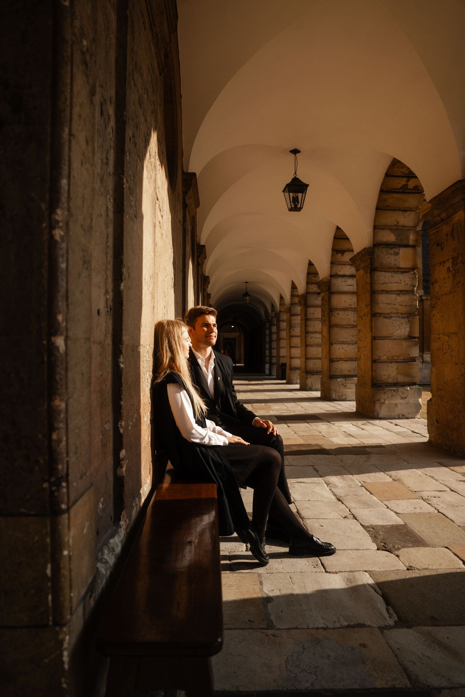 Couple in dark formal clothing sit on a wooden bench in a sunlit stone arcade with arched columns and a hanging lantern.