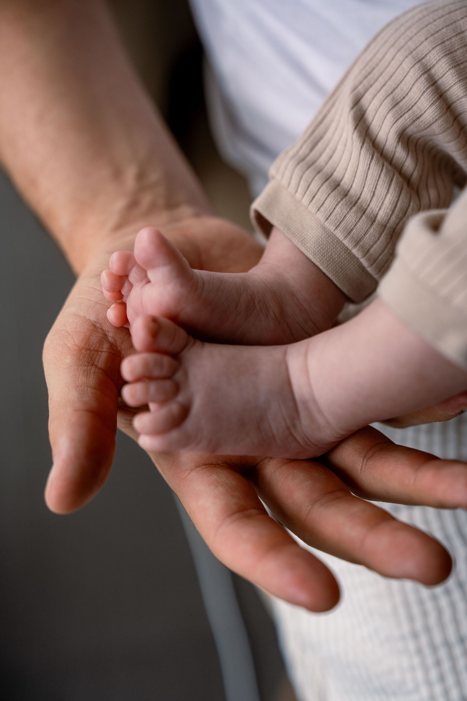 Adult hands gently cradling the tiny bare feet of a newborn baby dressed in a soft beige ribbed outfit.