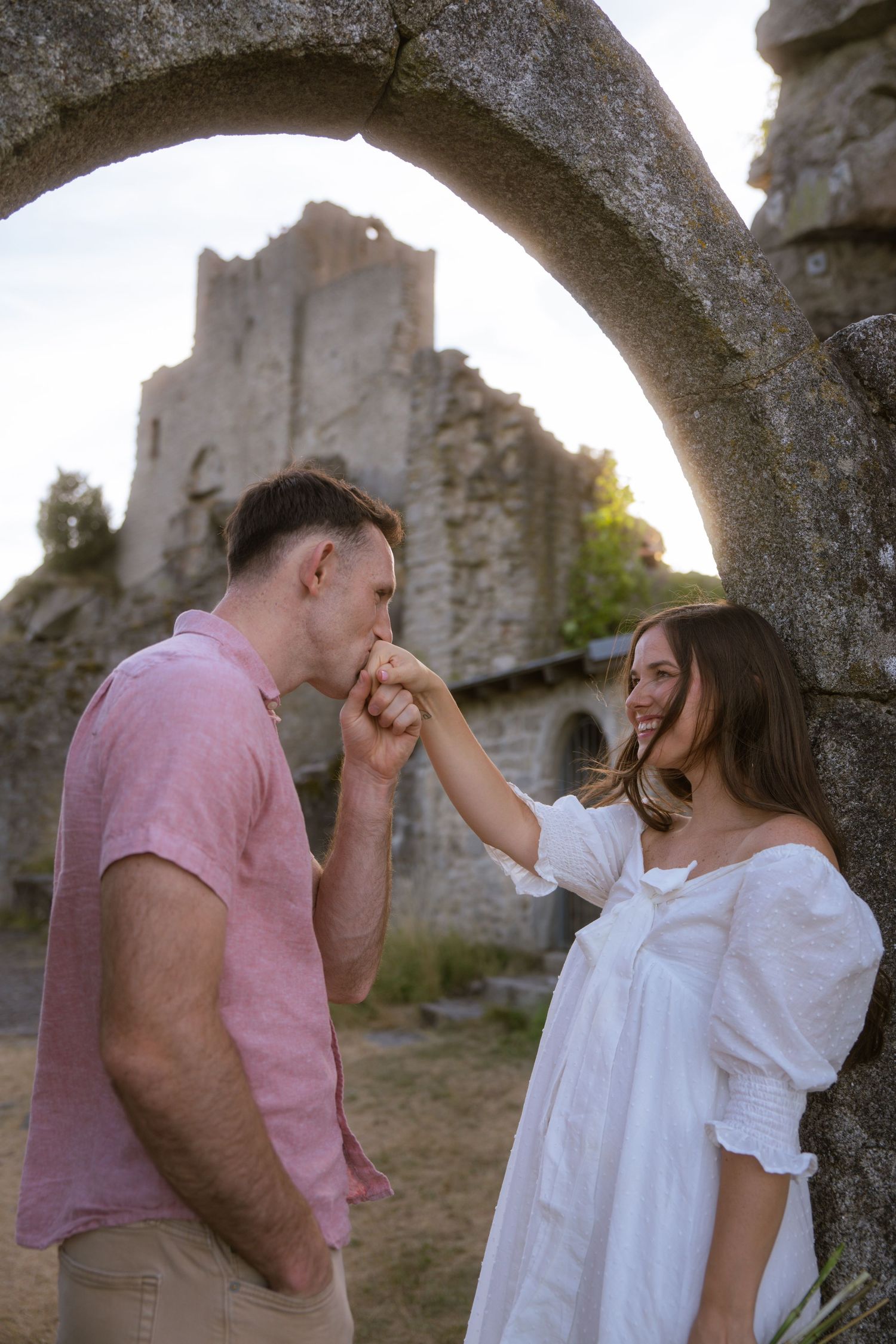 Man in a pink shirt kisses the hand of a smiling woman in a white dress beneath a stone arch with medieval castle ruins behind them.