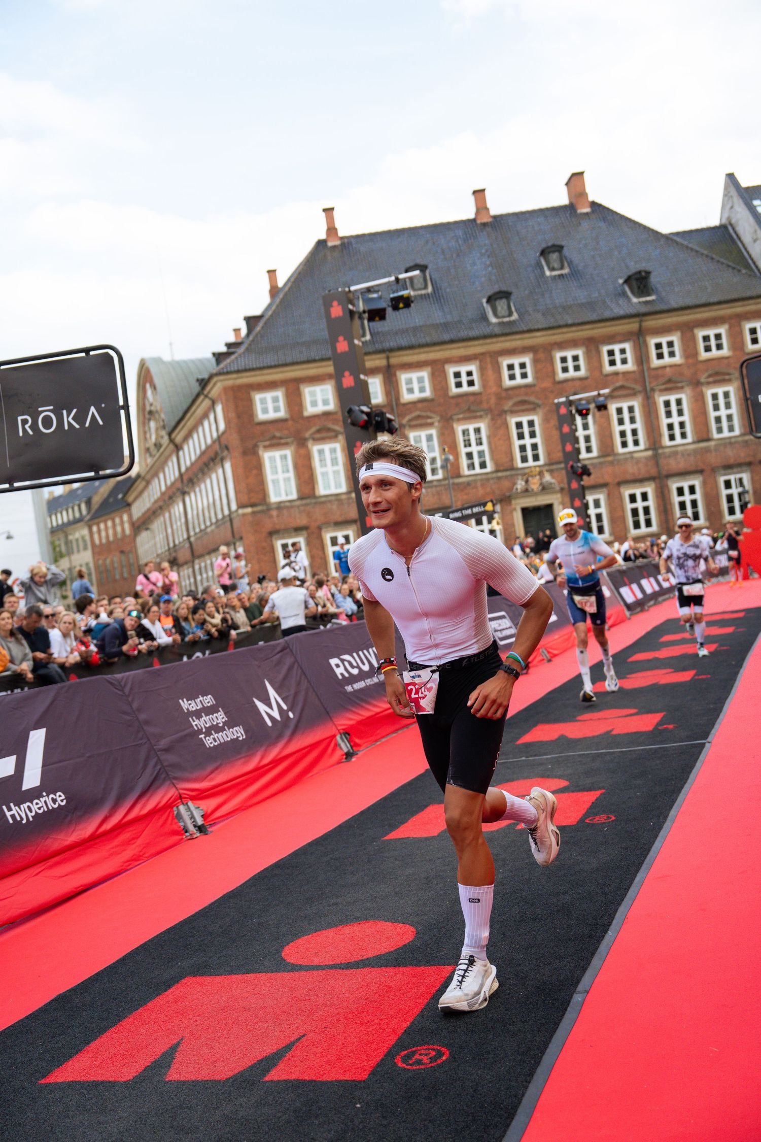 Triathlete in a white tri-suit and headband runs down a red Ironman finish carpet past cheering spectators and historic brick buildings.