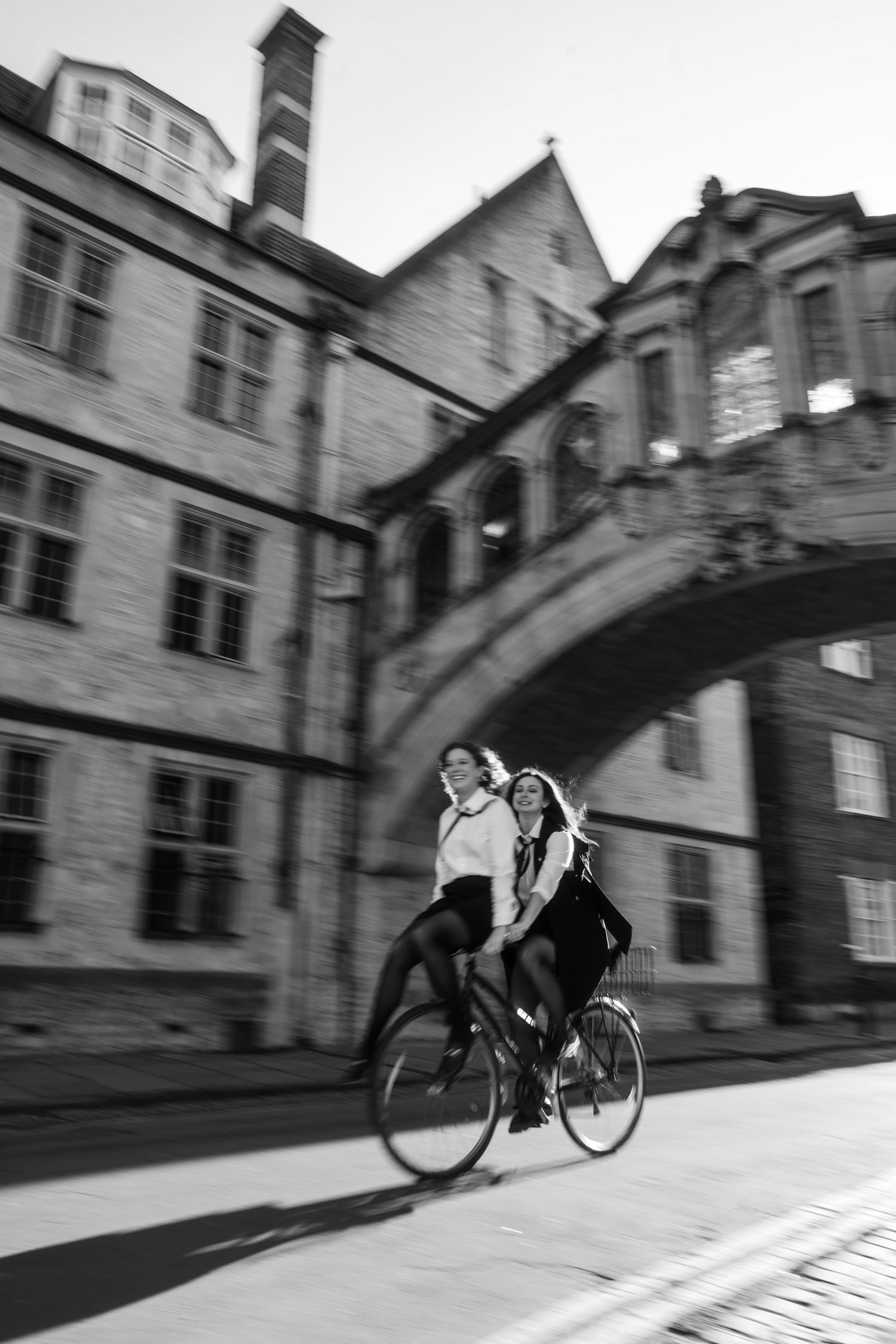 Two women ride a bicycle together past historic stone buildings and an arched covered bridge in a black and white motion-blur photo.
