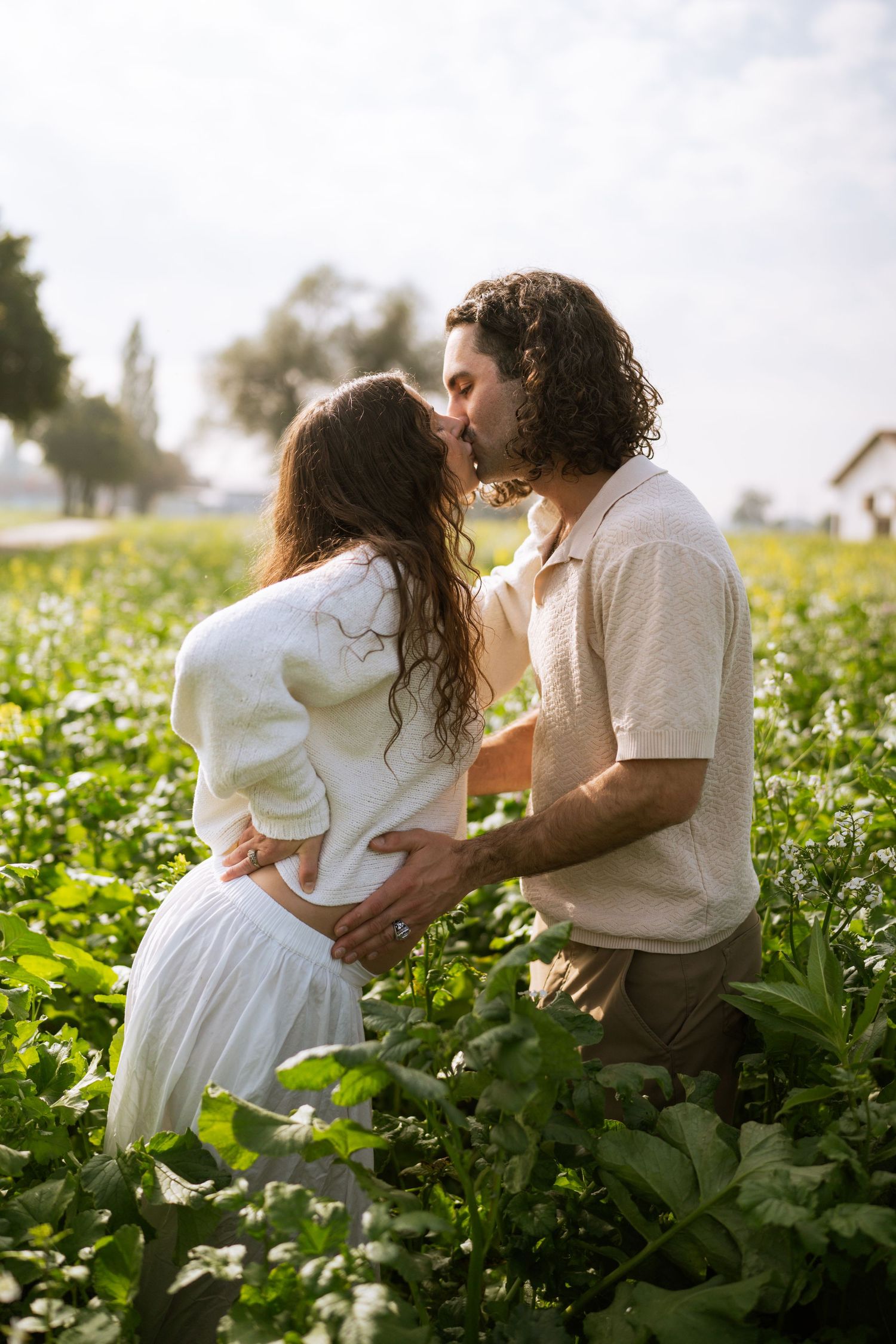 Expecting couple shares a kiss in a lush green field, the man's hand resting gently on her baby bump.