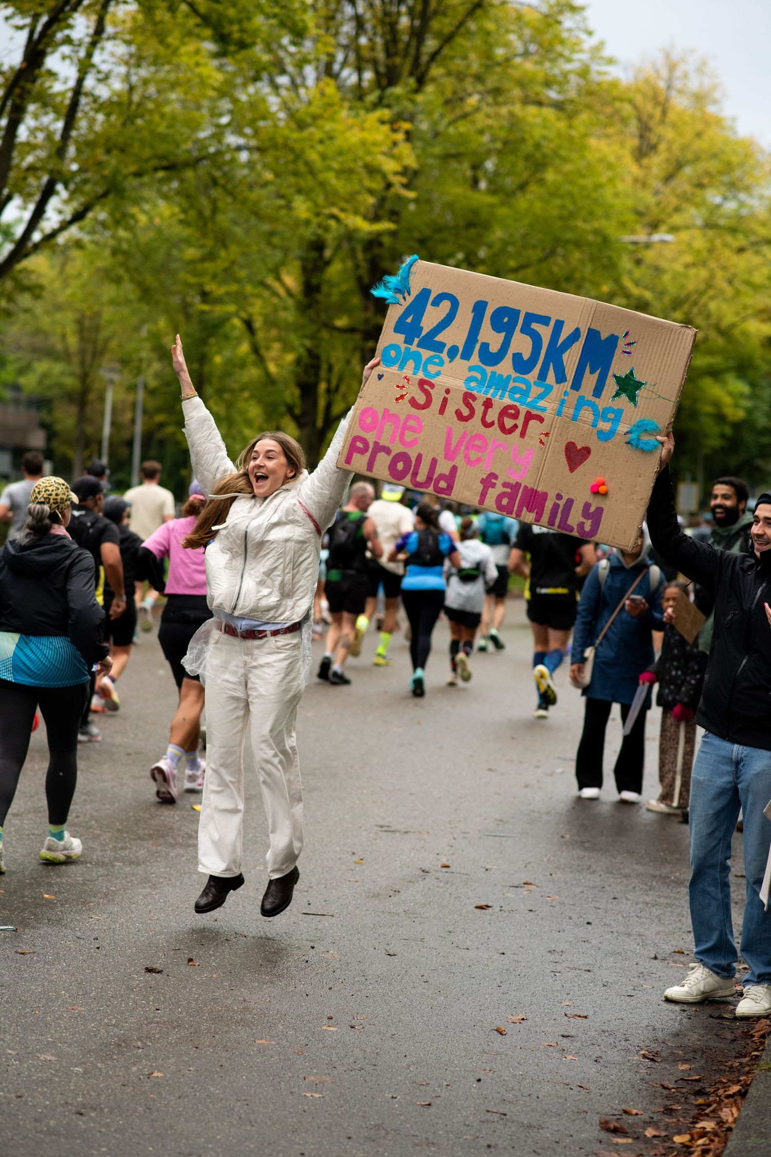 A woman in white jumps joyfully on a marathon course holding a sign cheering on her sister during the 42.195km race.