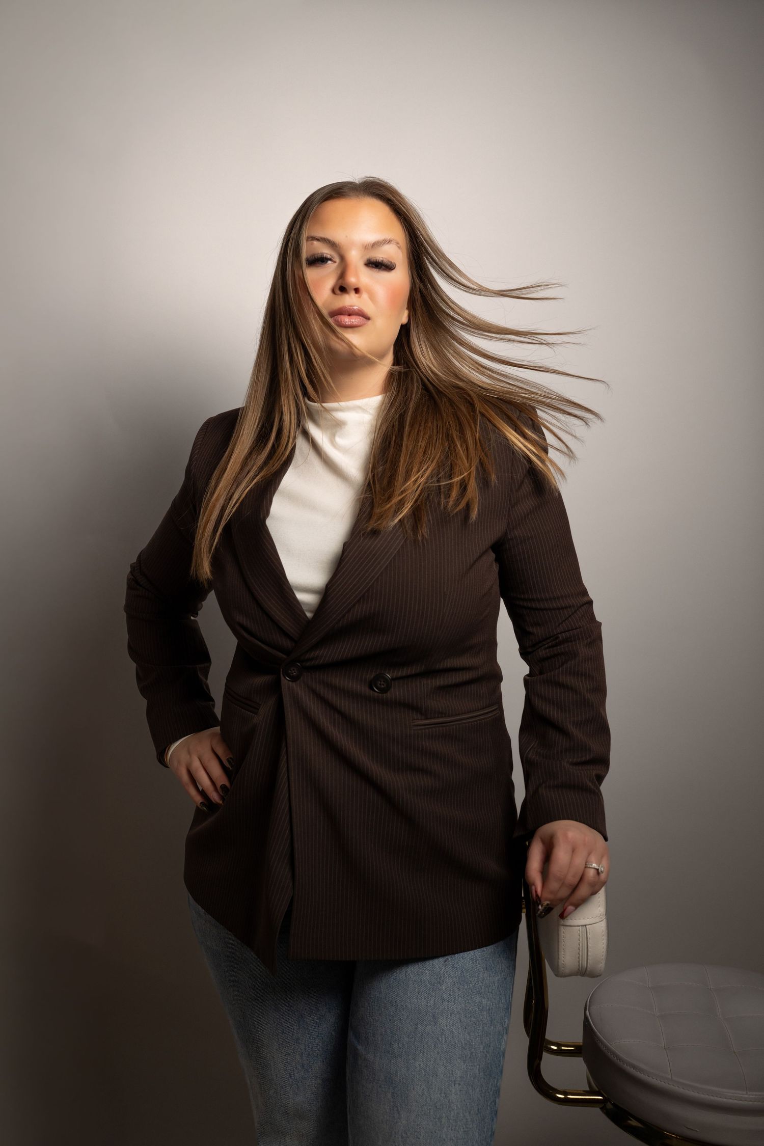 A woman in a dark brown blazer and jeans poses against a gray studio backdrop with her long hair flowing in the air.