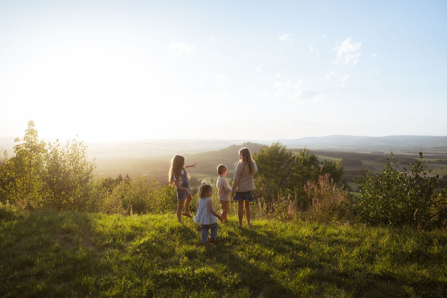 Four children standing on a grassy hilltop at sunset, looking out over a hazy countryside valley.