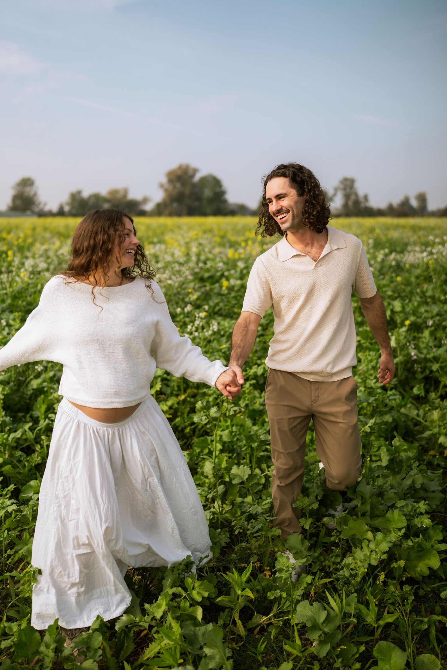 Smiling couple holds hands and walks through a yellow-flowered field, the pregnant woman in a white sweater and flowing skirt.