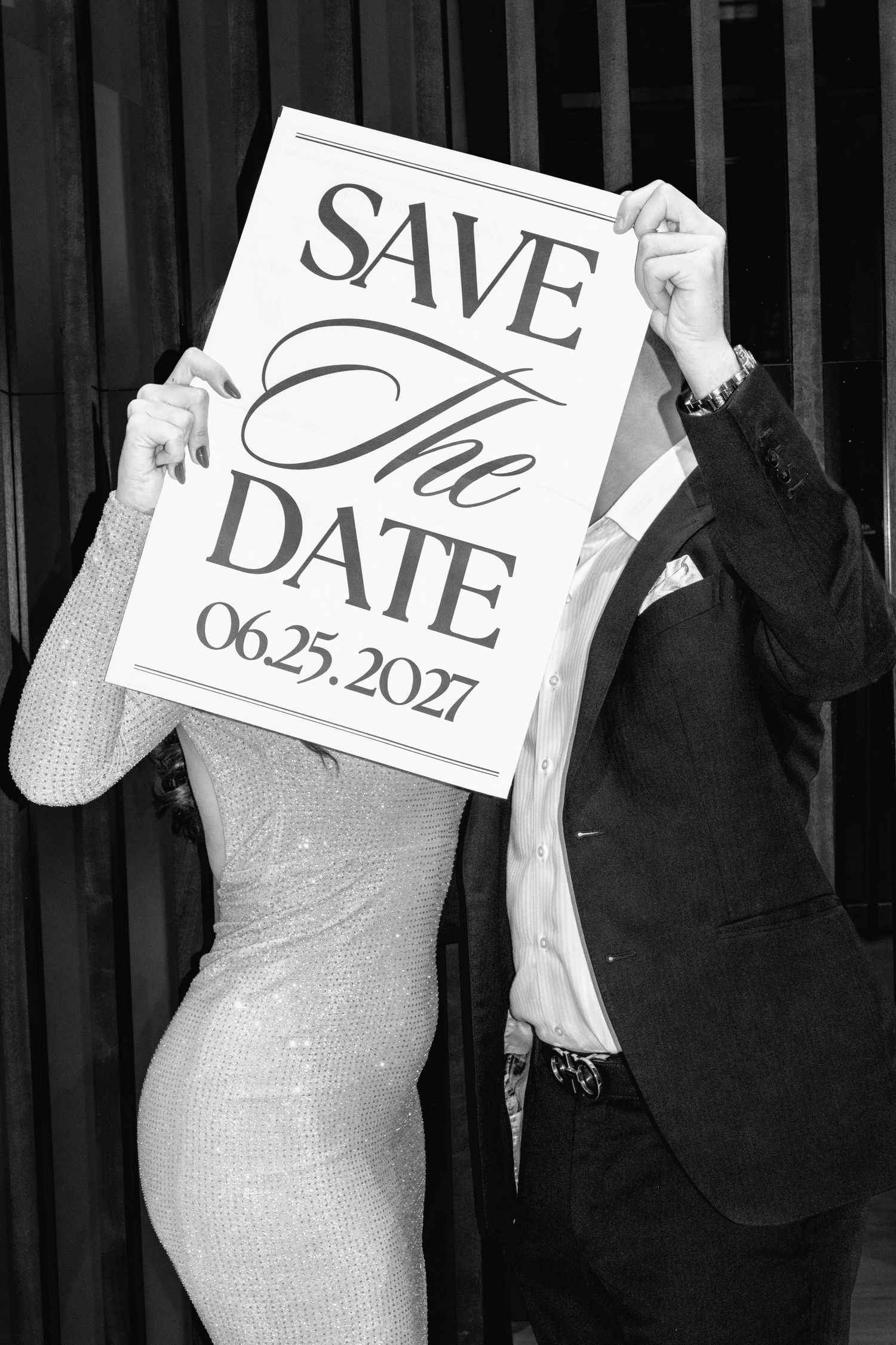 Black and white photo of a couple hiding their faces behind a Save the Date sign reading 06.25.2027.