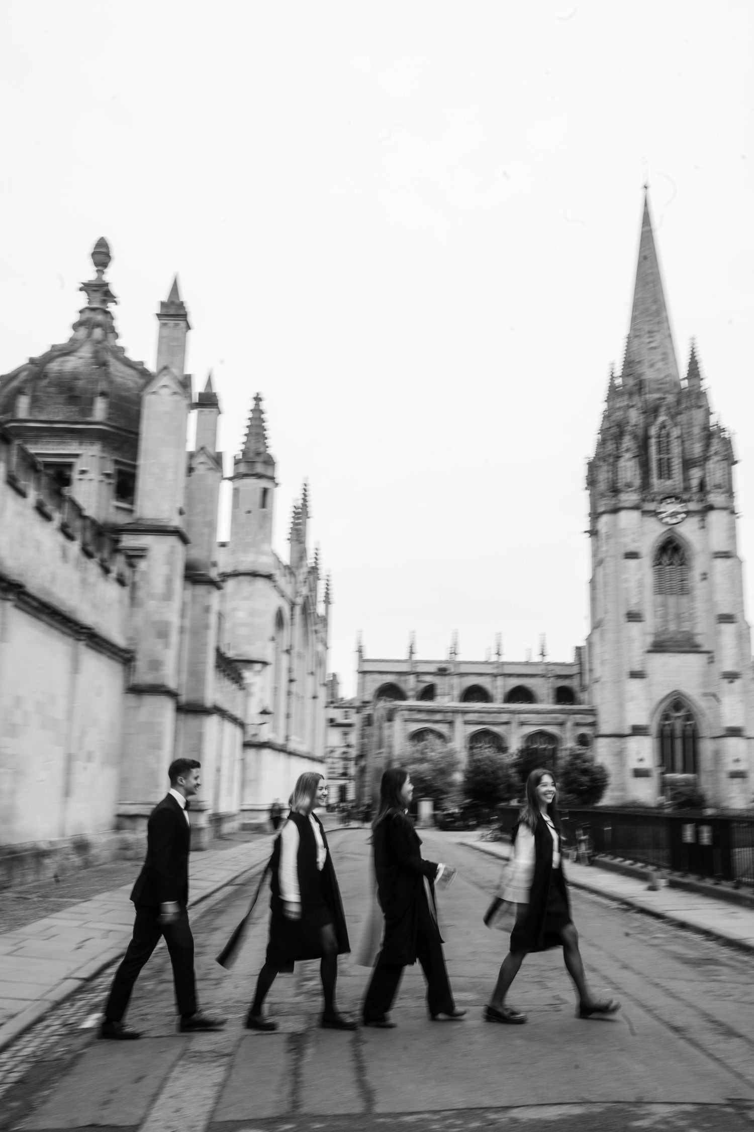 Four people in dark academic attire walk across a street in Oxford with Gothic spires and historic stone buildings behind them.