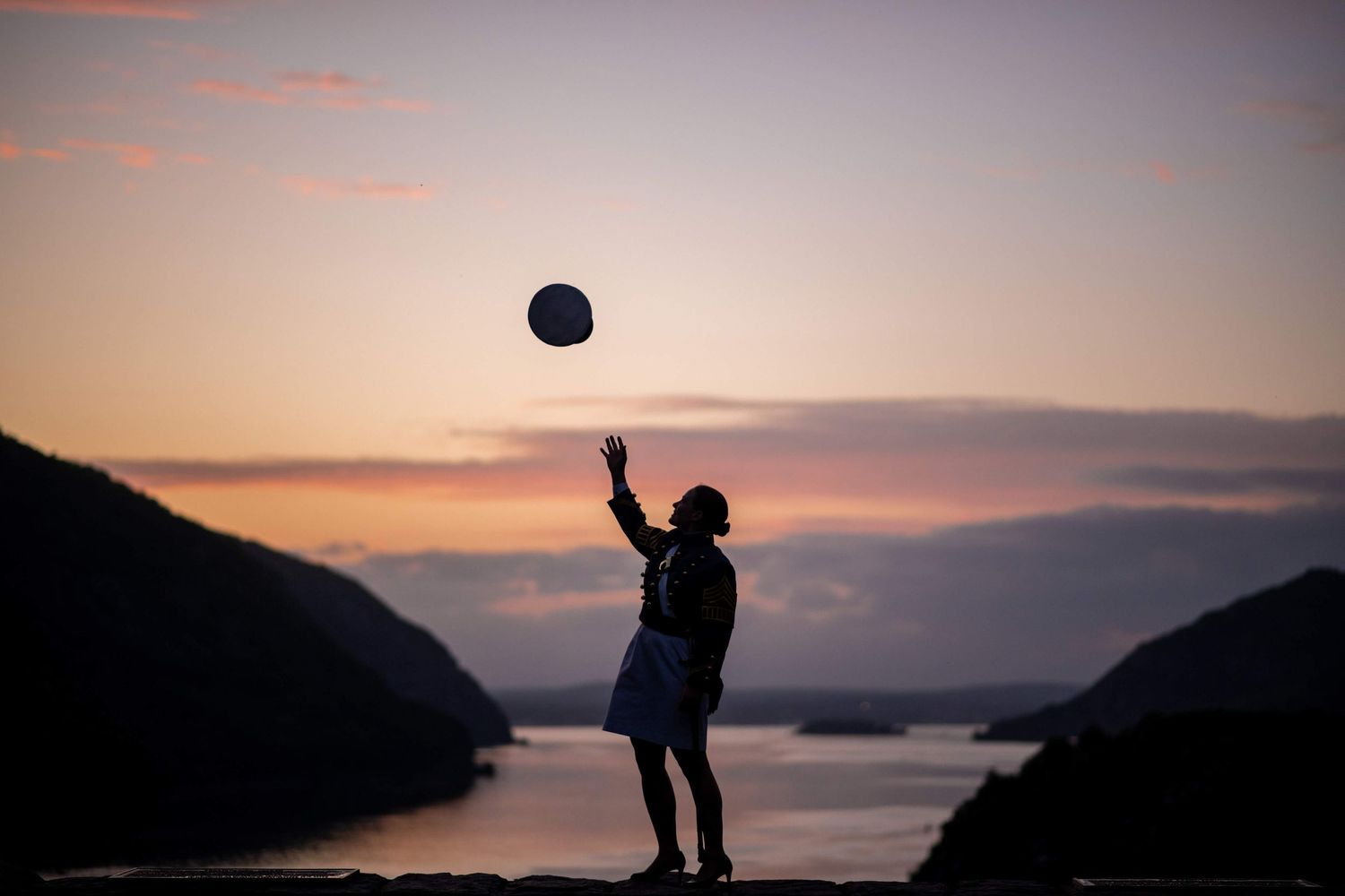 A silhouetted person tosses a ball into the air against a colorful sunset sky above hills and a calm bay.
