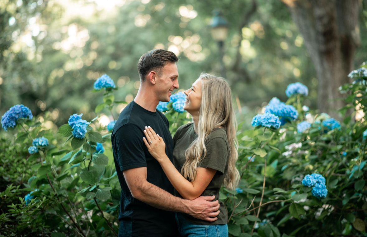 A smiling couple embraces among blooming blue hydrangeas in a sunlit garden.