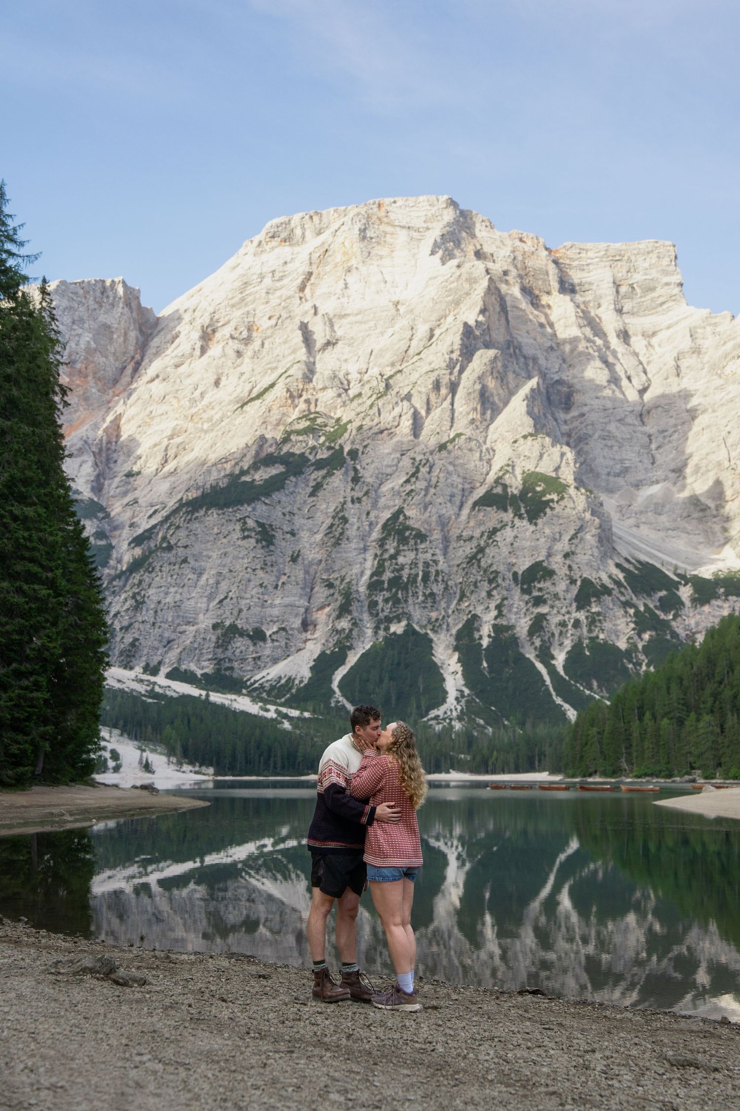 Couple kissing on the pebbly shore of an alpine lake with a towering rocky mountain and evergreen forest reflected in the calm water.
