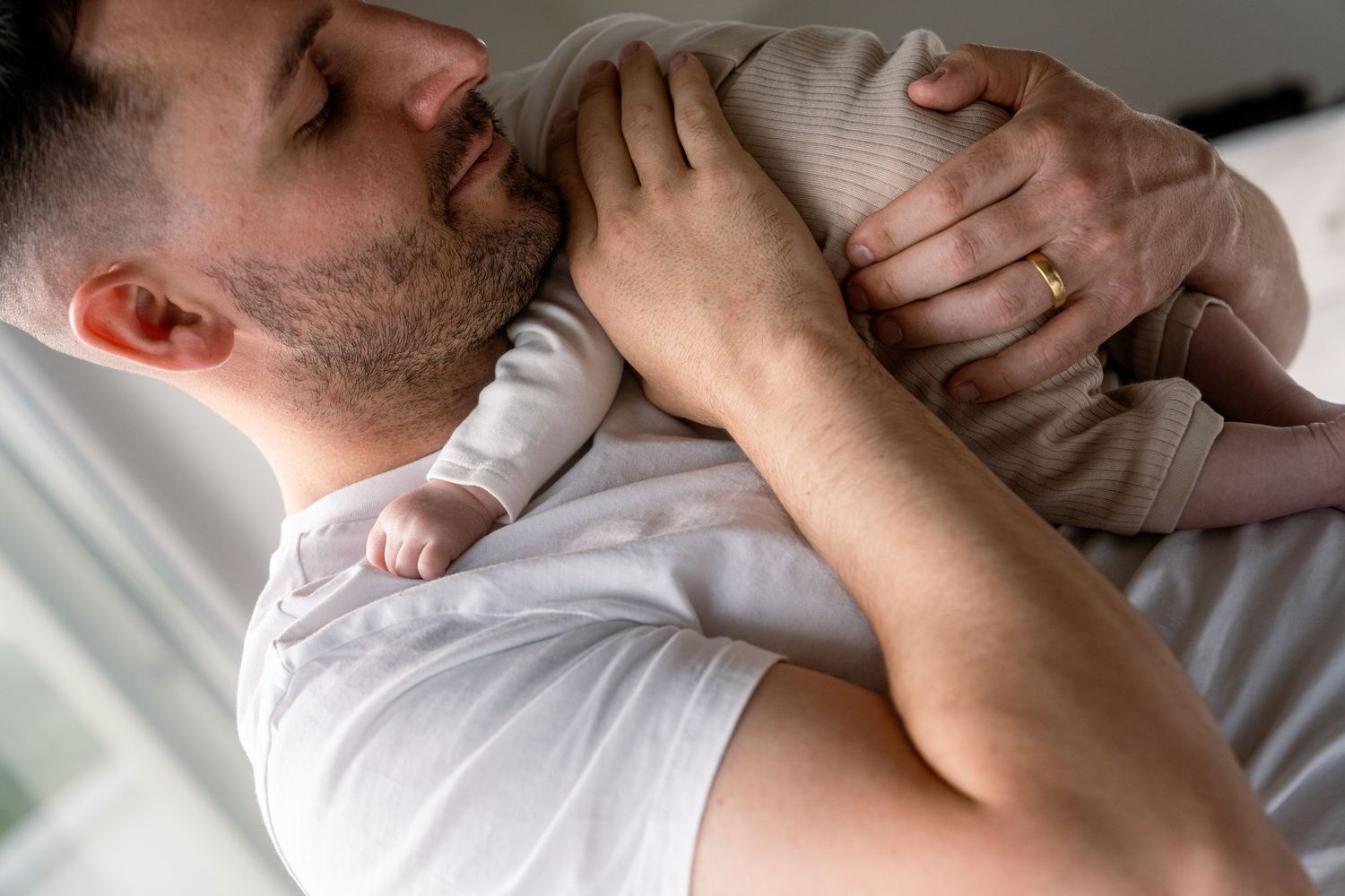 A bearded father in a white t-shirt gently cradles a sleeping newborn baby against his chest in a tender, quiet moment.