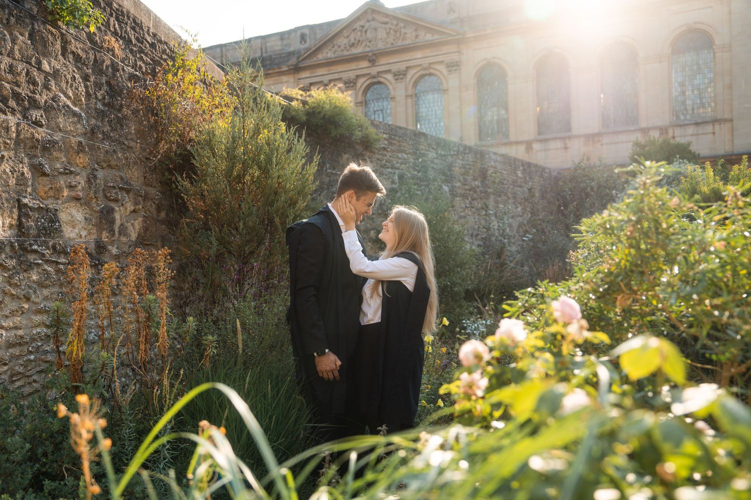 Couple embracing in a sunlit garden beside an old stone wall, with blooming flowers and a historic building glowing in the background.