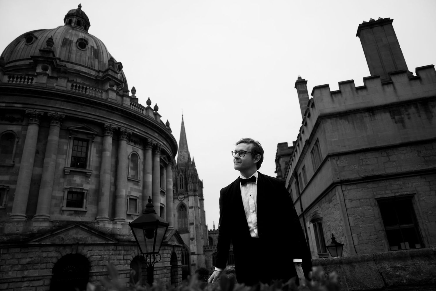 Man in a tuxedo and bow tie stands on a historic Oxford street beside the Radcliffe Camera dome in a black and white photo.