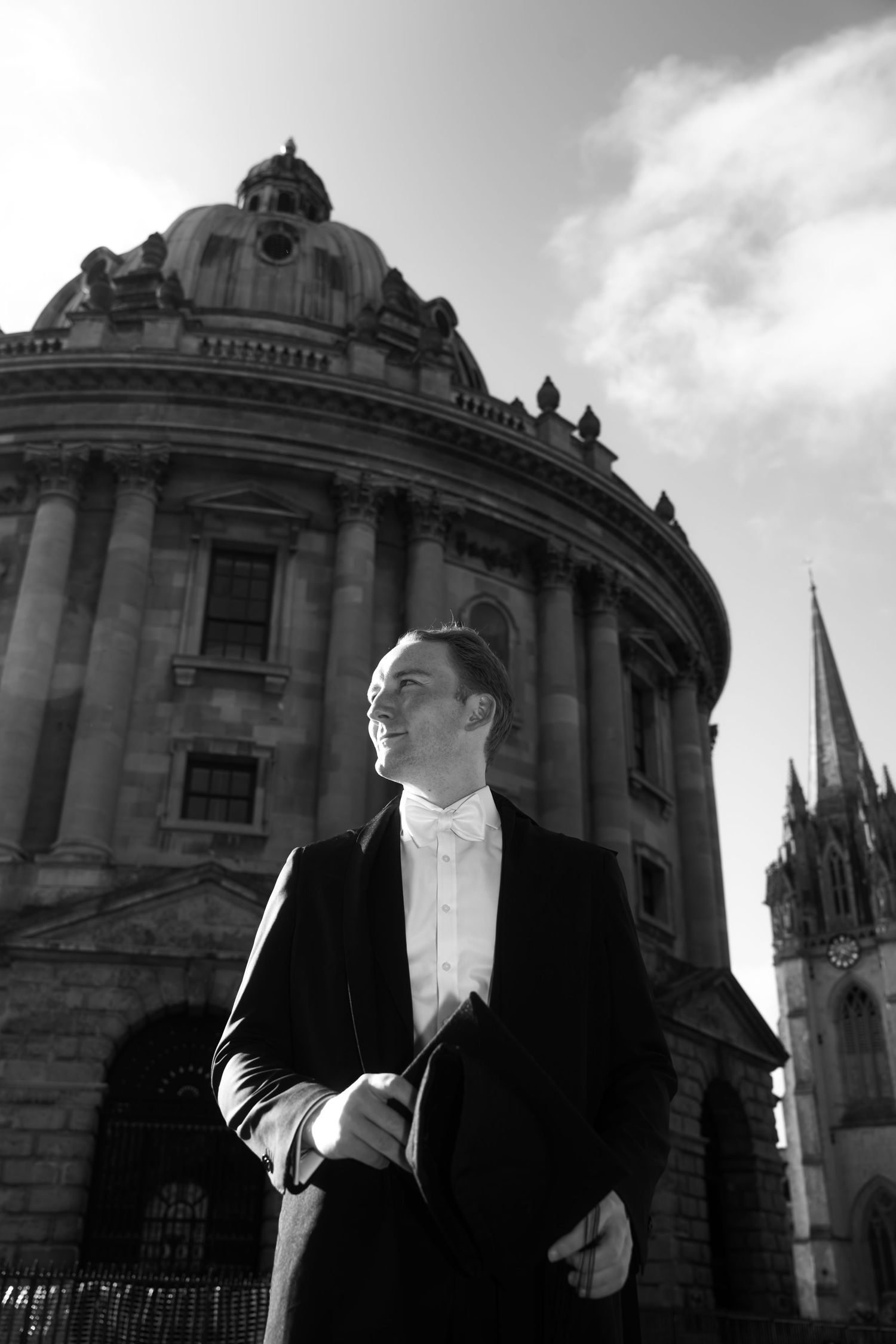 Man in formal black suit and white bow tie holds a mortarboard before the Radcliffe Camera and a church spire in Oxford, in black and white.