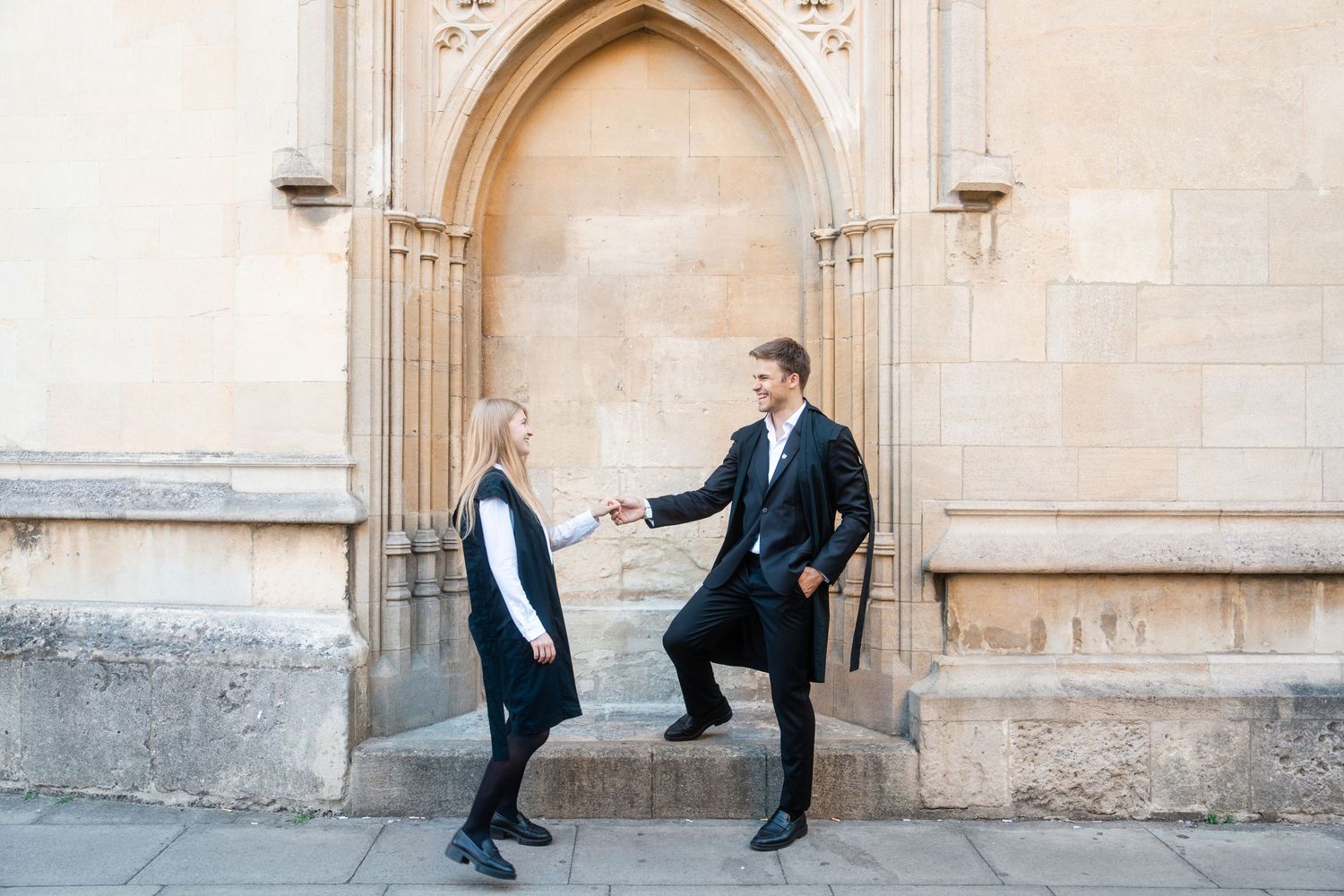 Couple in dark formal outfits holding hands in front of an arched stone doorway on a historic building.