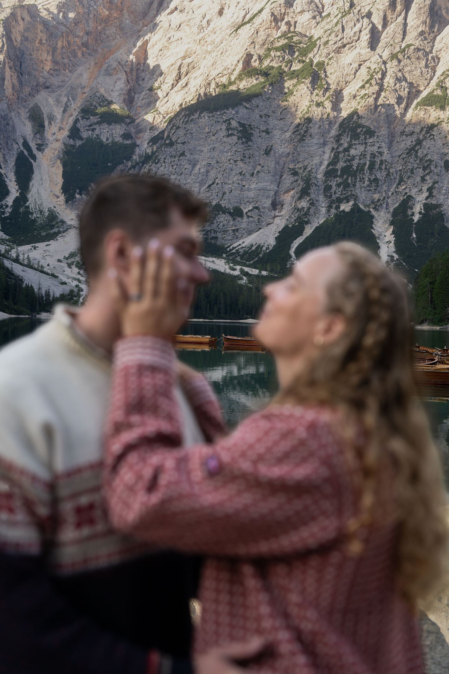Couple embracing in a romantic moment by a mountain lake, the woman in a red knit sweater touching his face, with rocky Dolomites behind.