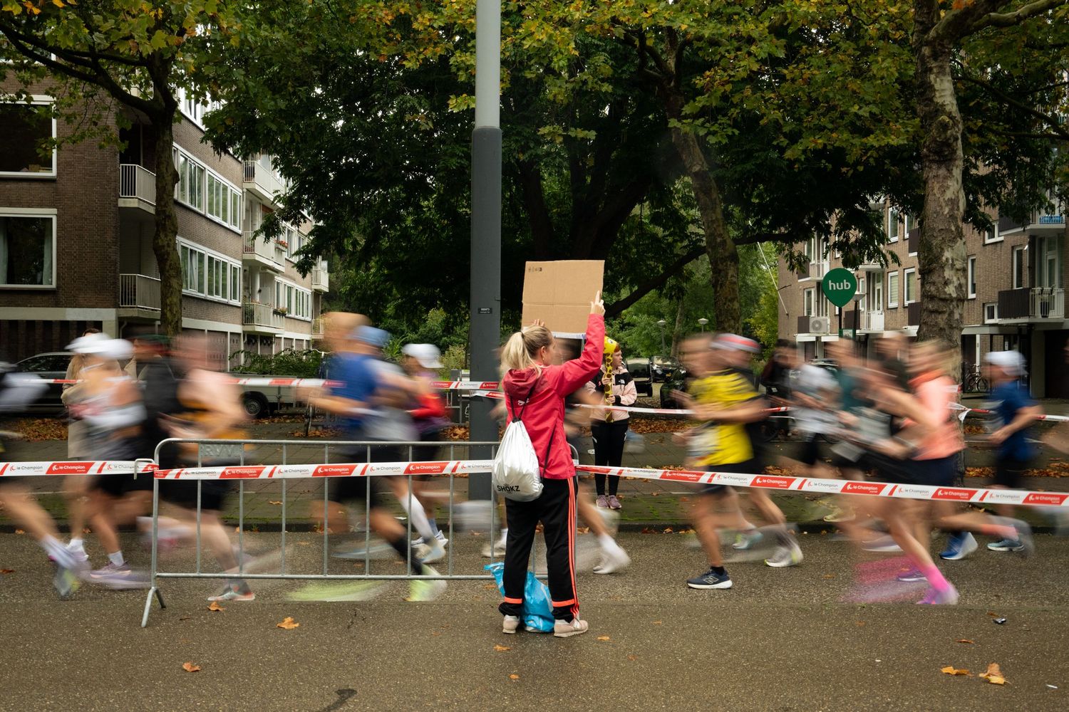 Spectator in a red jacket holds a cardboard sign above a barrier as blurred marathon runners race past on a tree-lined street.