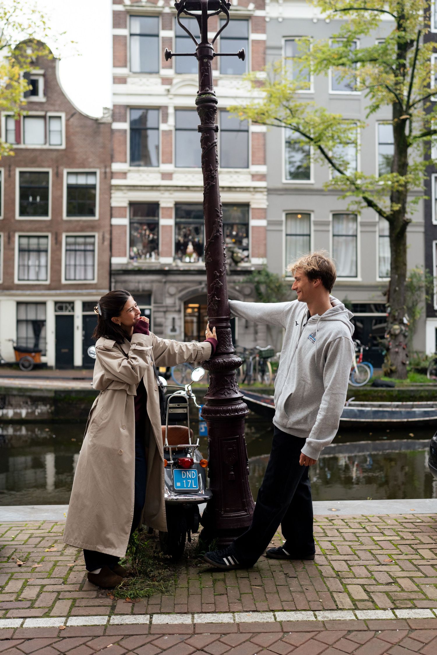 Woman in a trench coat and smiling man in a hoodie playfully leaning on a lamppost by an Amsterdam canal.