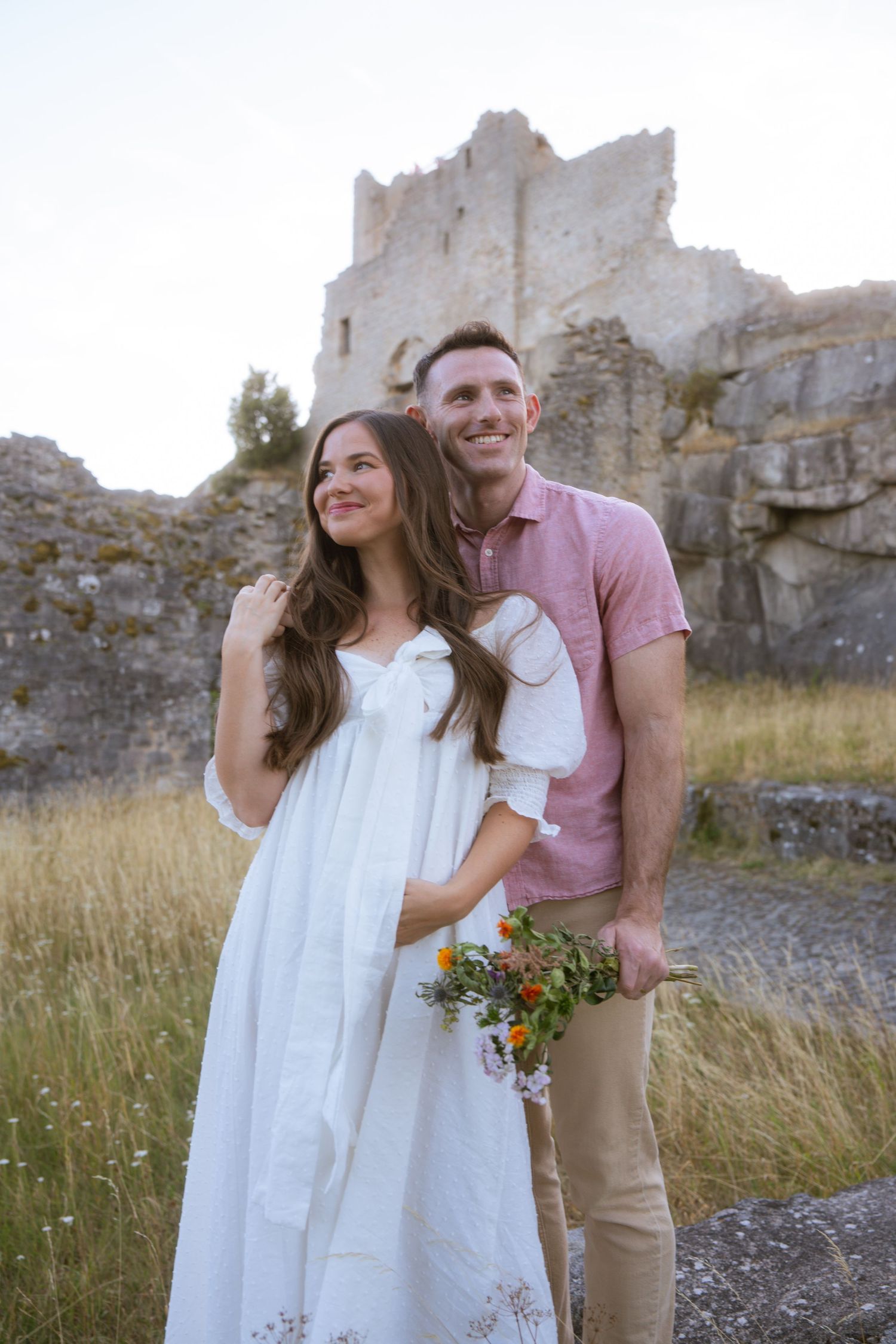 Smiling couple embracing in front of ancient stone castle ruins, the woman in a white dress holding a wildflower bouquet in a grassy field.