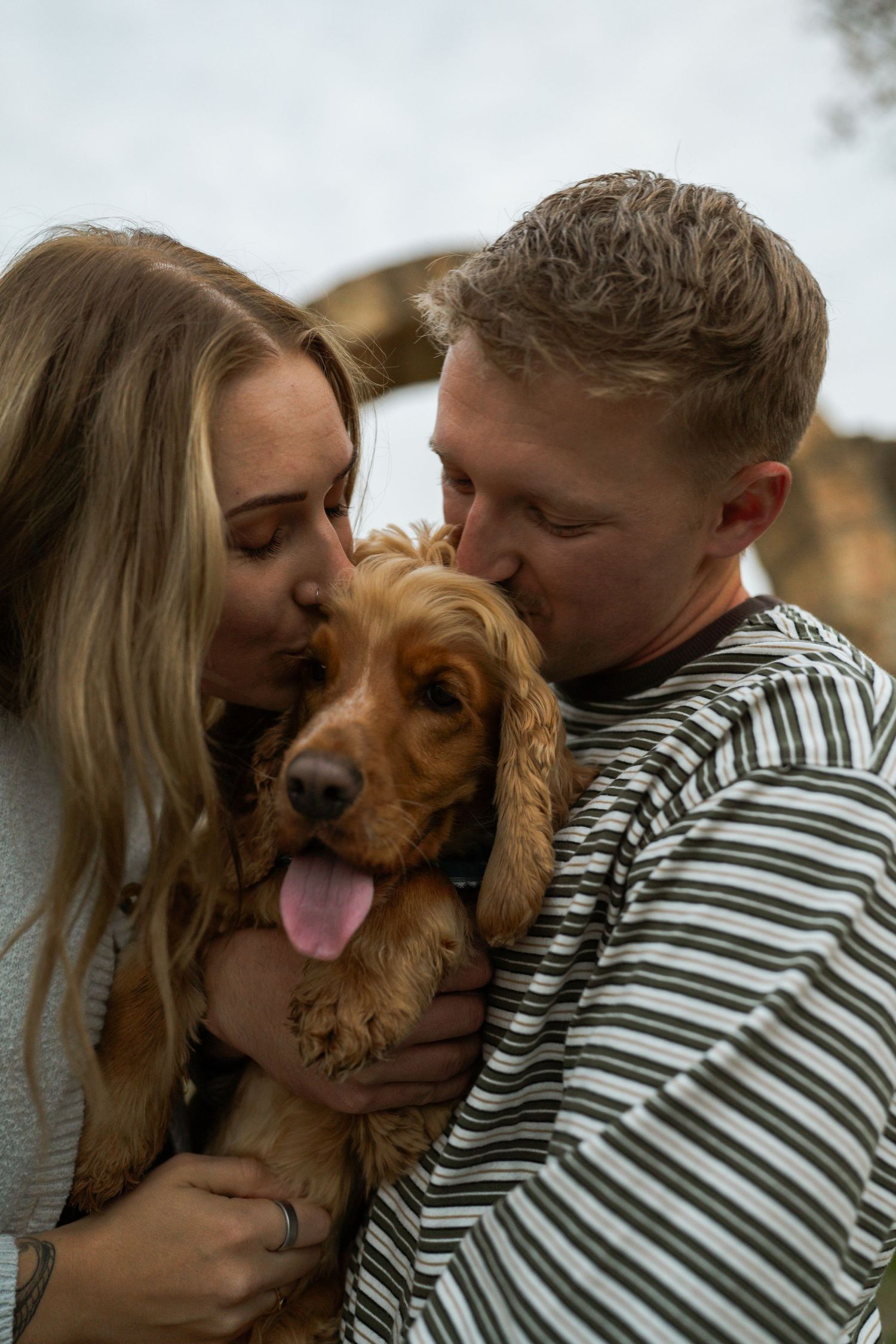 Young couple affectionately kissing their golden cocker spaniel dog, which smiles with its tongue out.