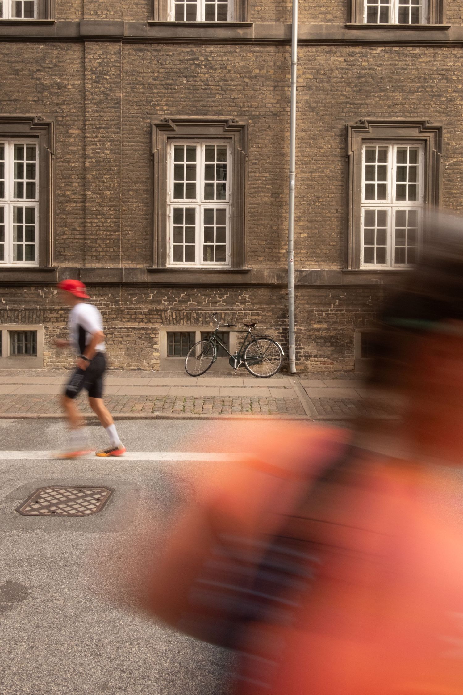 Runner in a red cap jogs past a brick building and a parked bicycle on a city street, with a blurred figure in the foreground.