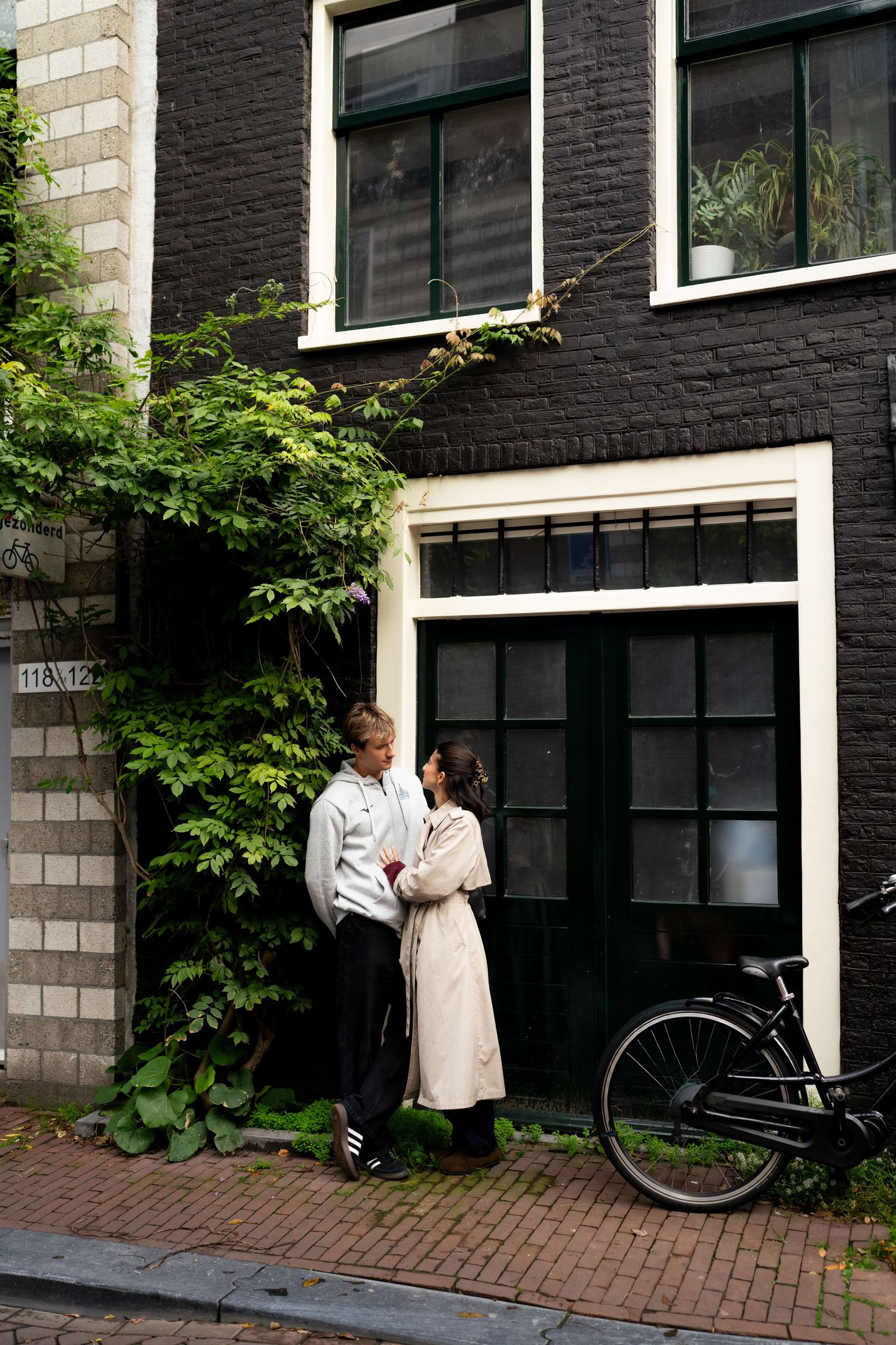 Couple embracing beside a leafy vine-covered brick facade, with a bicycle parked near a dark green door.