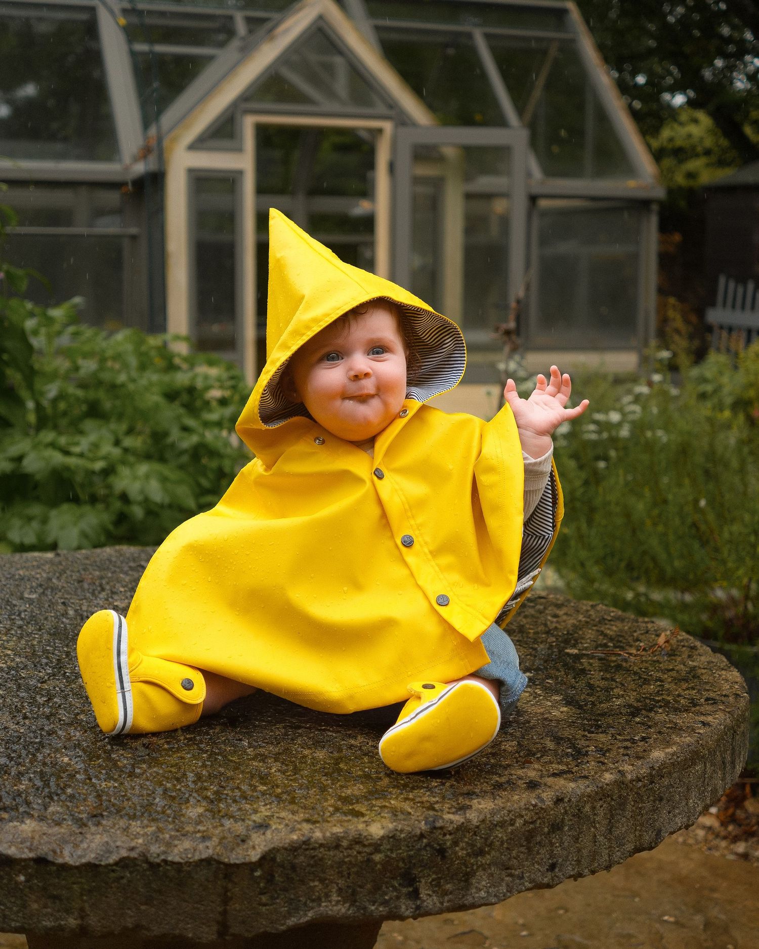 Baby in a yellow hooded rain poncho and matching boots sits on a stone garden table, waving, with a greenhouse and plants behind.