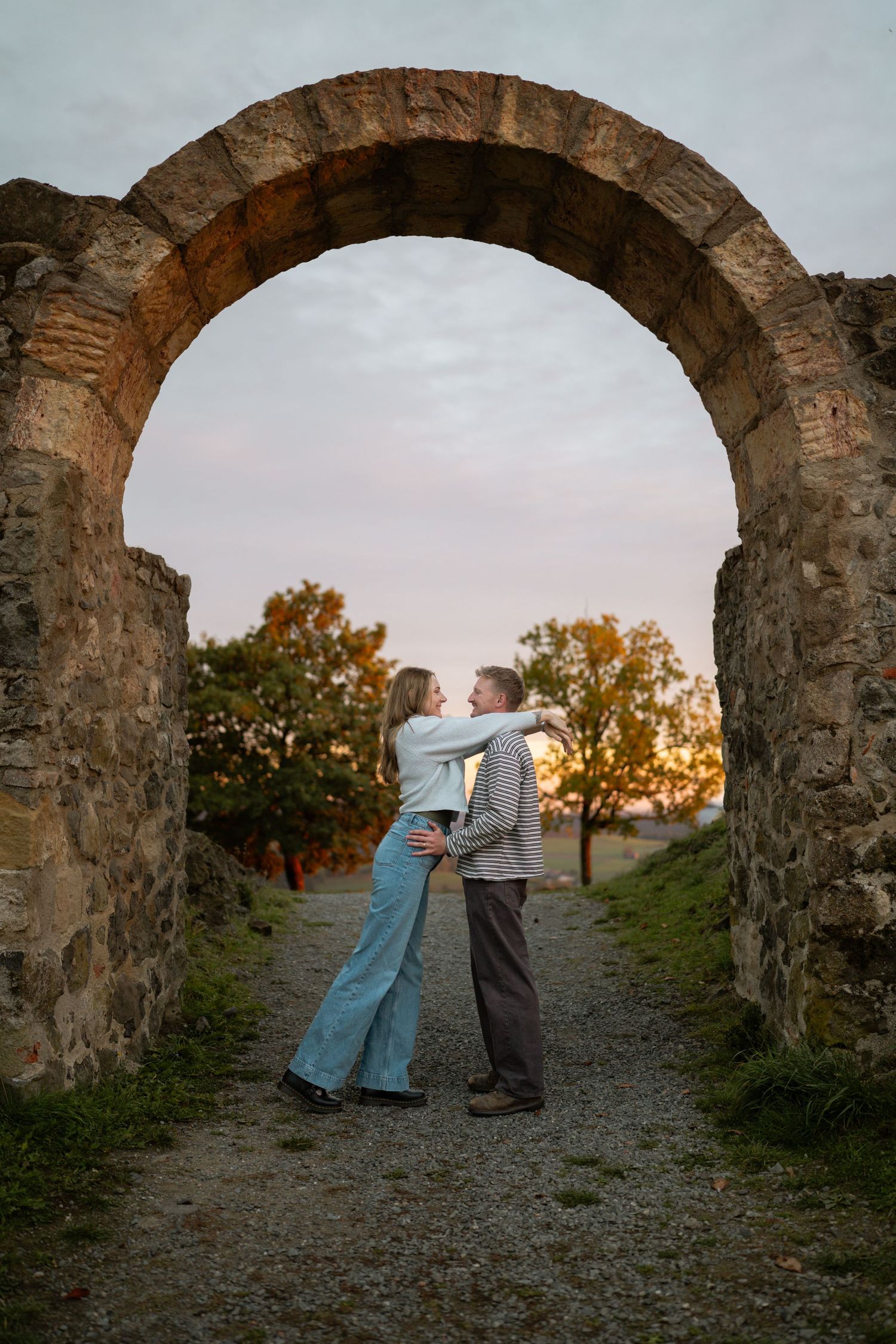 Couple embracing beneath a rustic stone archway on a gravel path at sunset with autumn trees behind them.