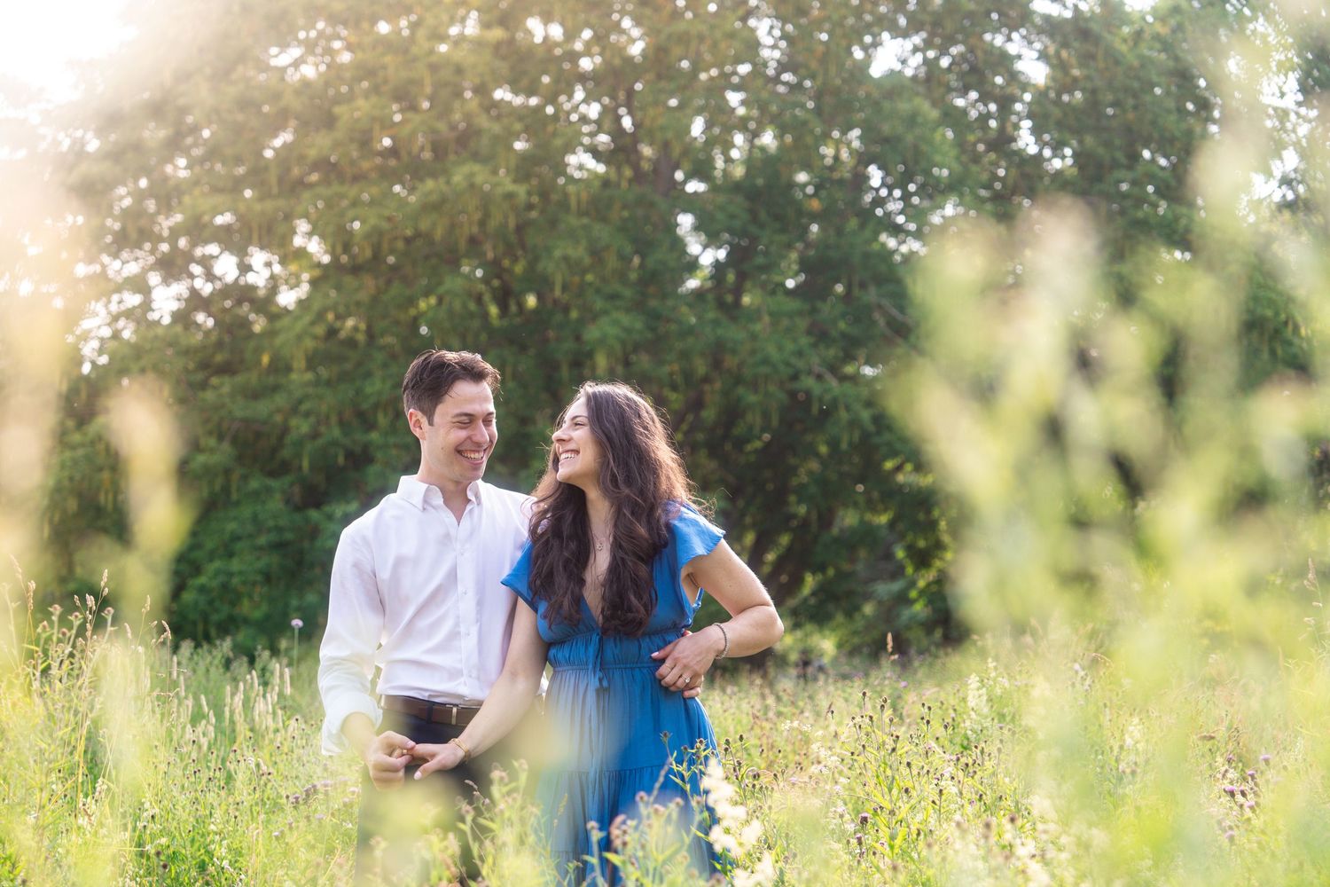 Smiling couple embrace in a sunlit grassy meadow with trees behind them, the woman in a blue dress and the man in a white shirt.