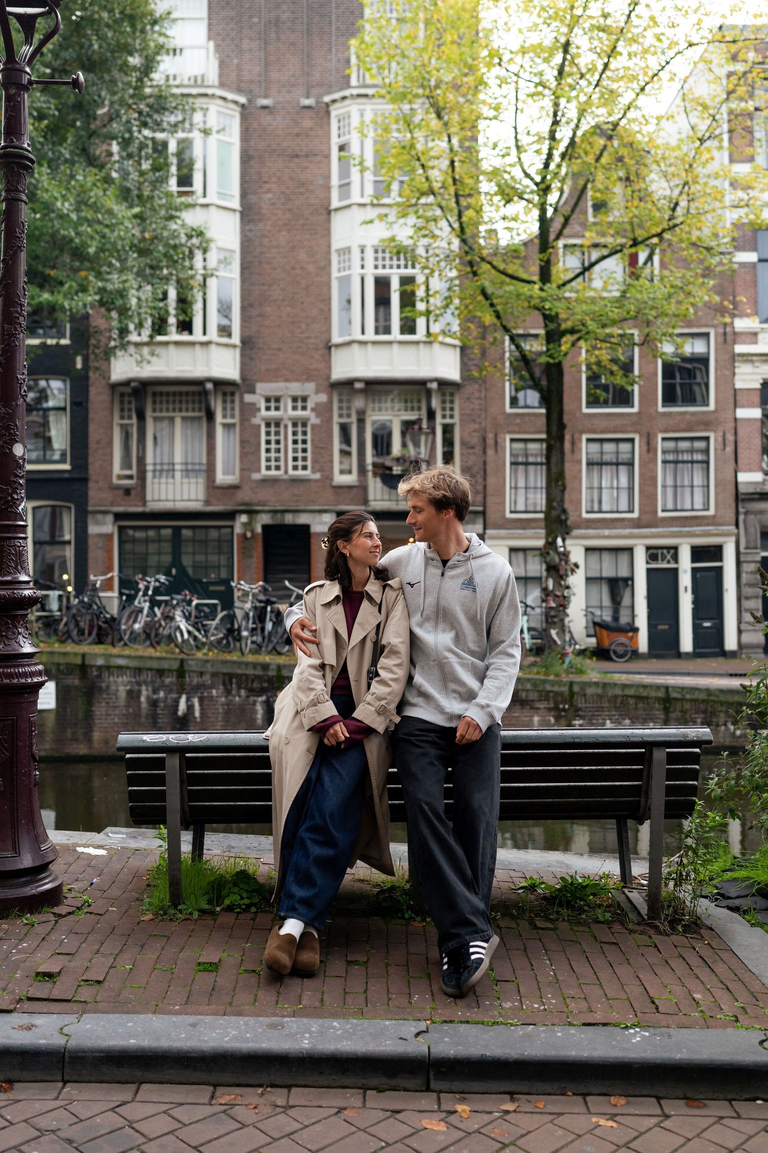 Couple sitting close together on a wooden bench along an Amsterdam canal with traditional brick townhouses behind.